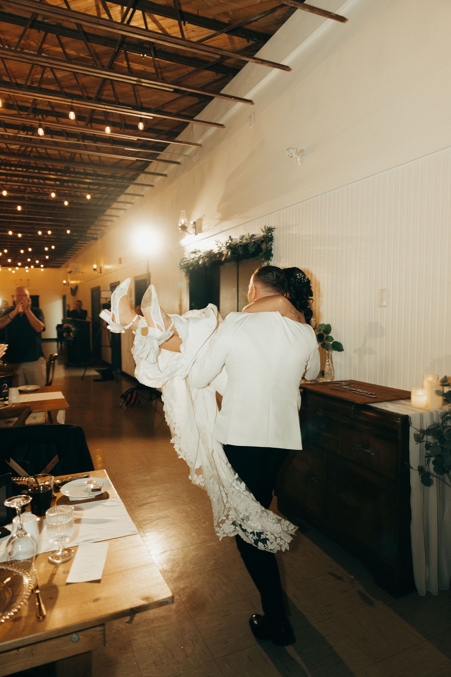 Wedding reception venue with rustic wooden tables and white decor in an industrial-style space with exposed ceiling beams.