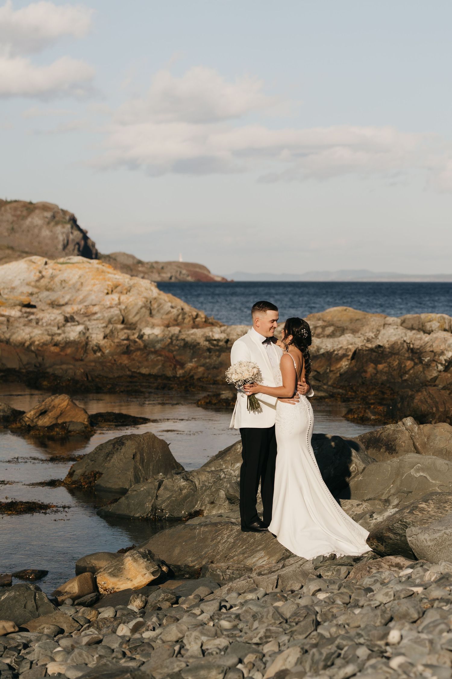 Couple in wedding attire embraces on rocky coastal shoreline with ocean and cliffs in background at sunset.