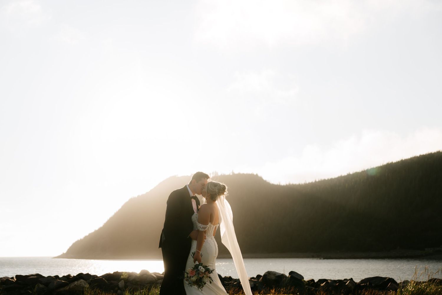 Bride and groom share a romantic kiss at sunset with ocean and lush green mountains in the background.