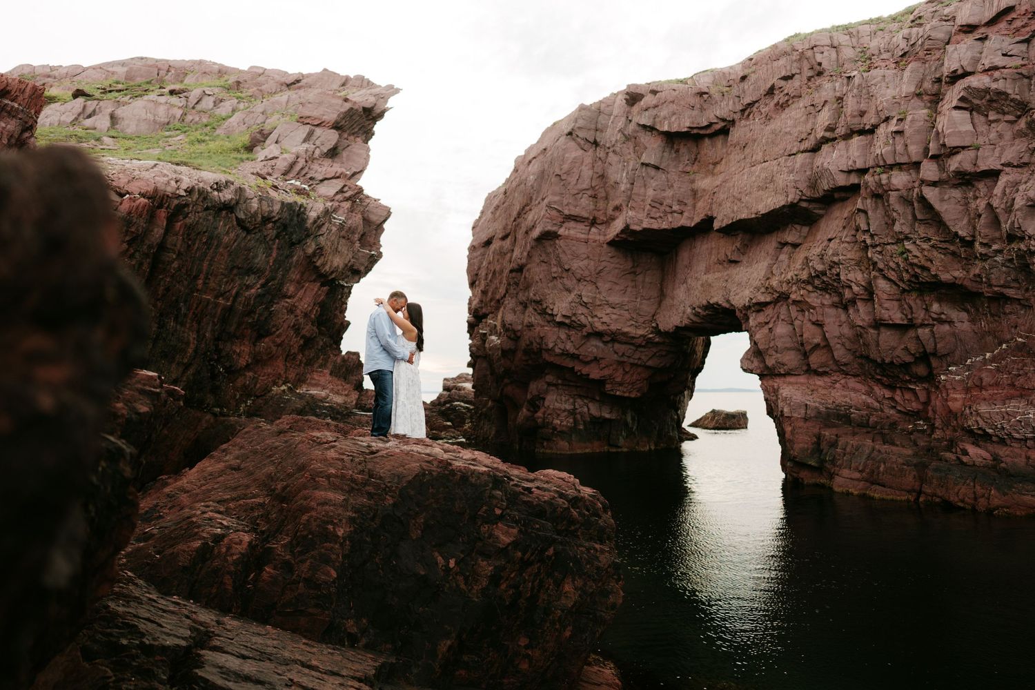 Couple embracing on dramatic red rock arch formation by the ocean, romantic coastal engagement photo.