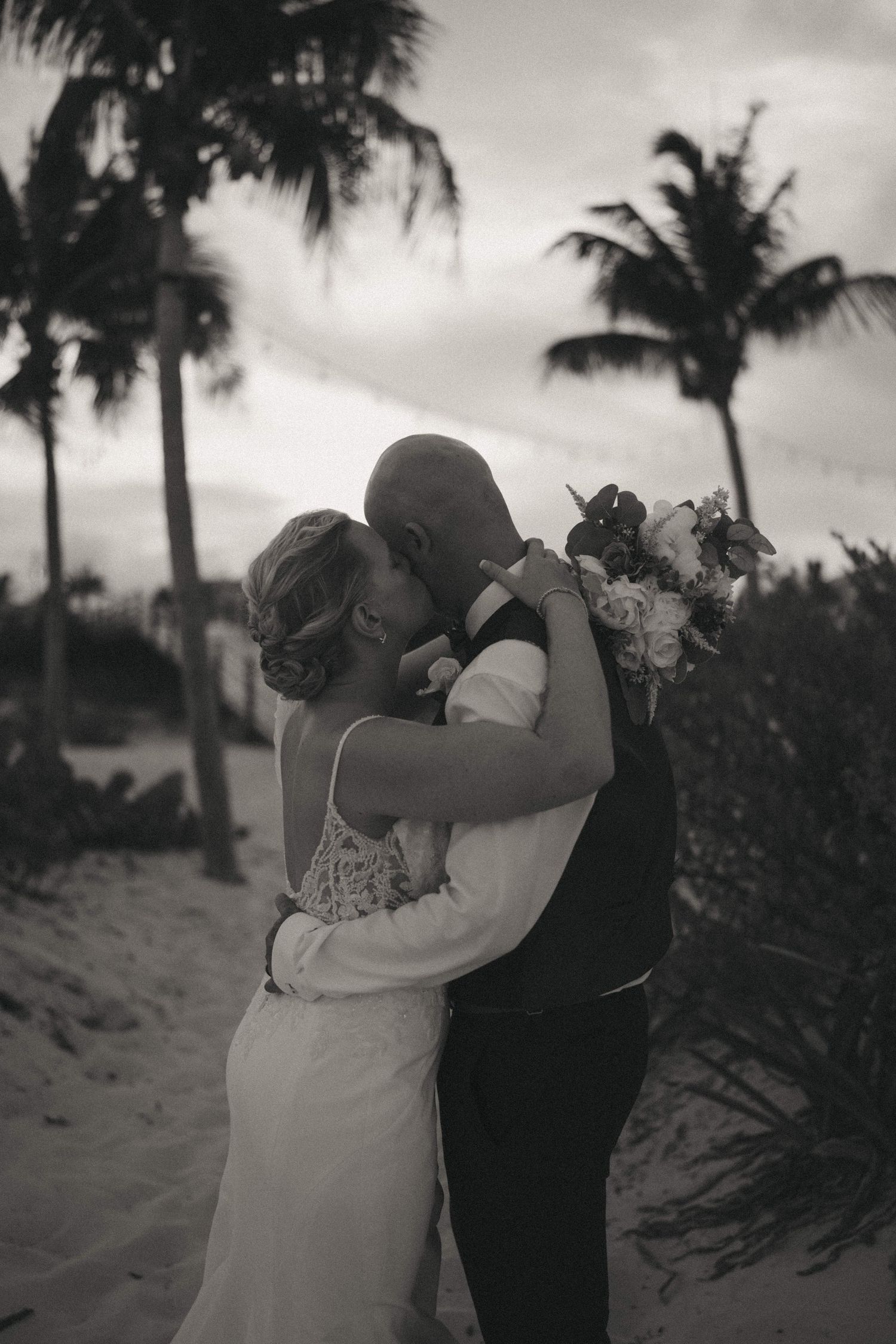 Black and white photo of newlyweds sharing an intimate moment on a palm-lined beach.