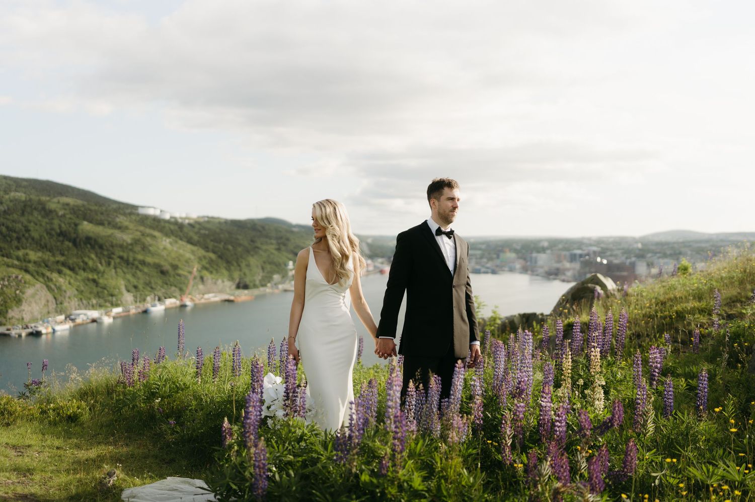 Wedding couple stands among purple lupine flowers with coastal view in background.