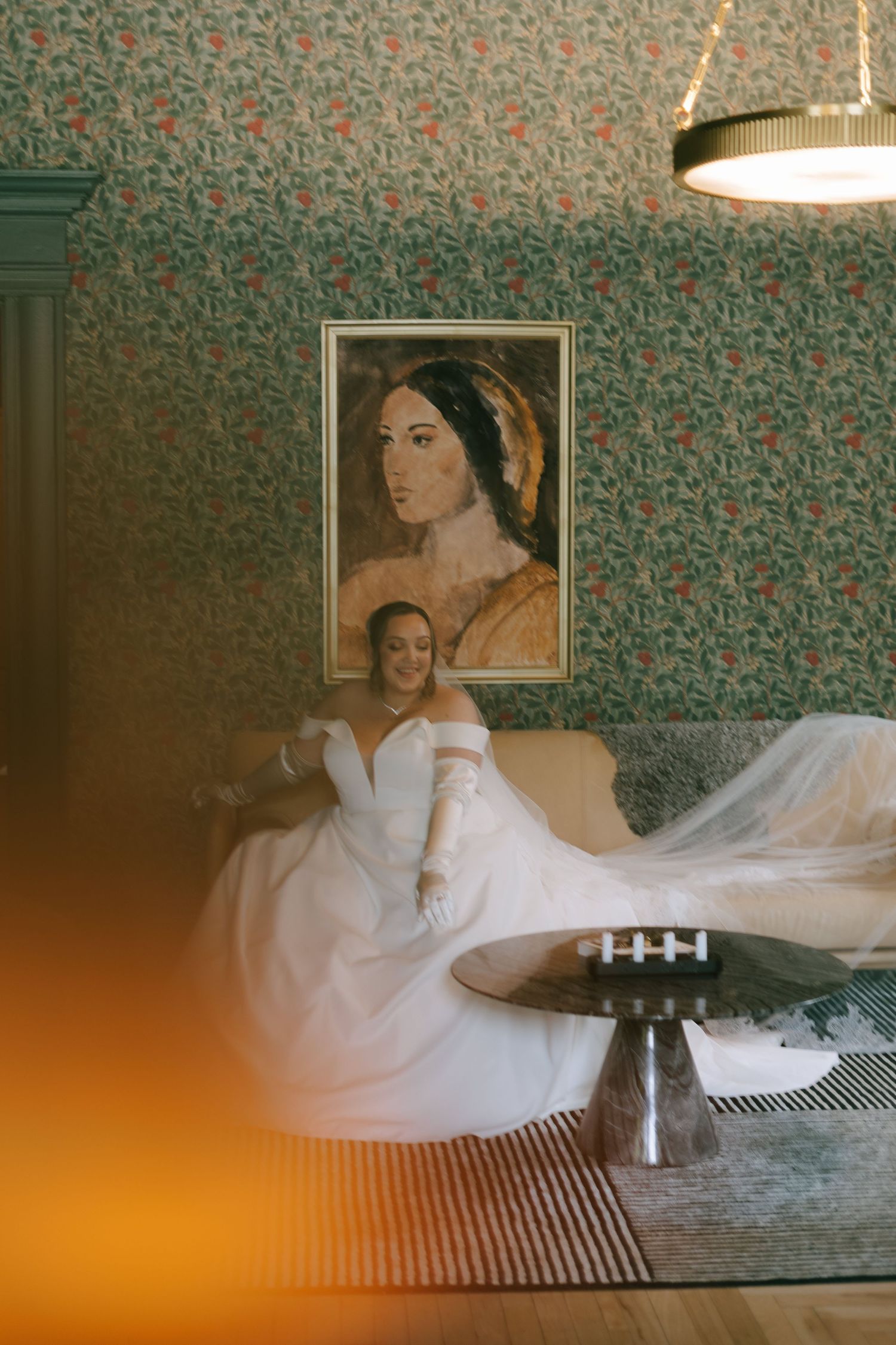 Bride in white dress reclining on vintage bed with floral wallpaper in moody lighting.