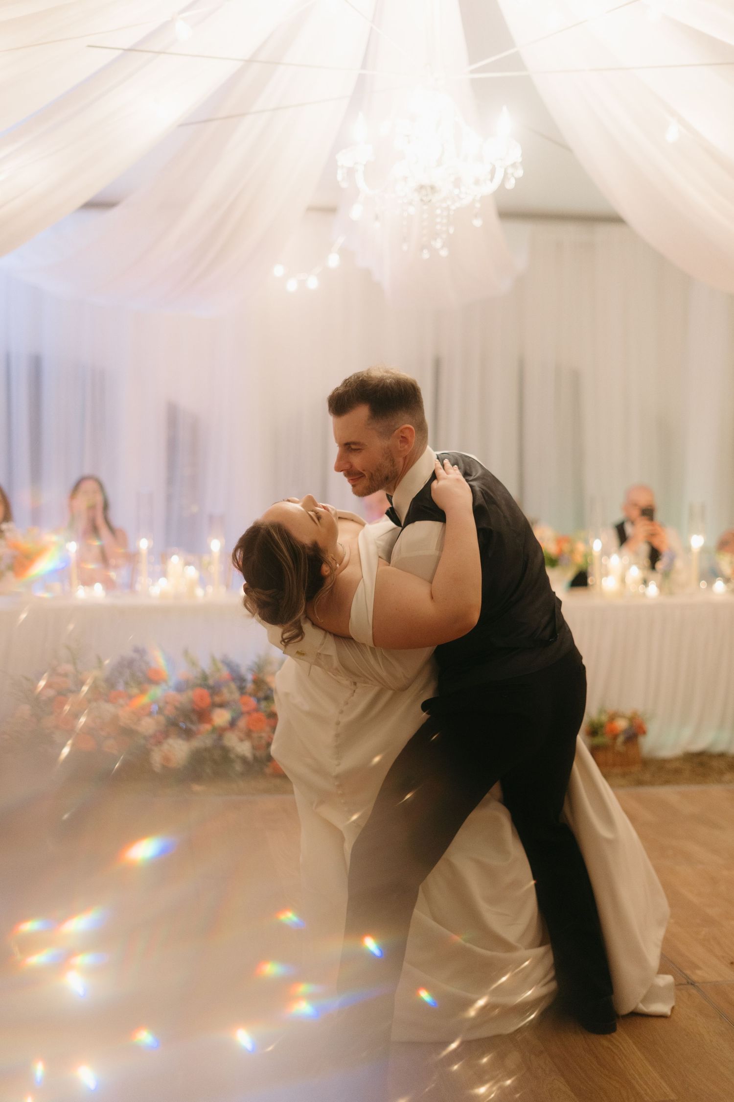 Newlyweds share an intimate dance moment under white tent draping with rainbow light reflections.