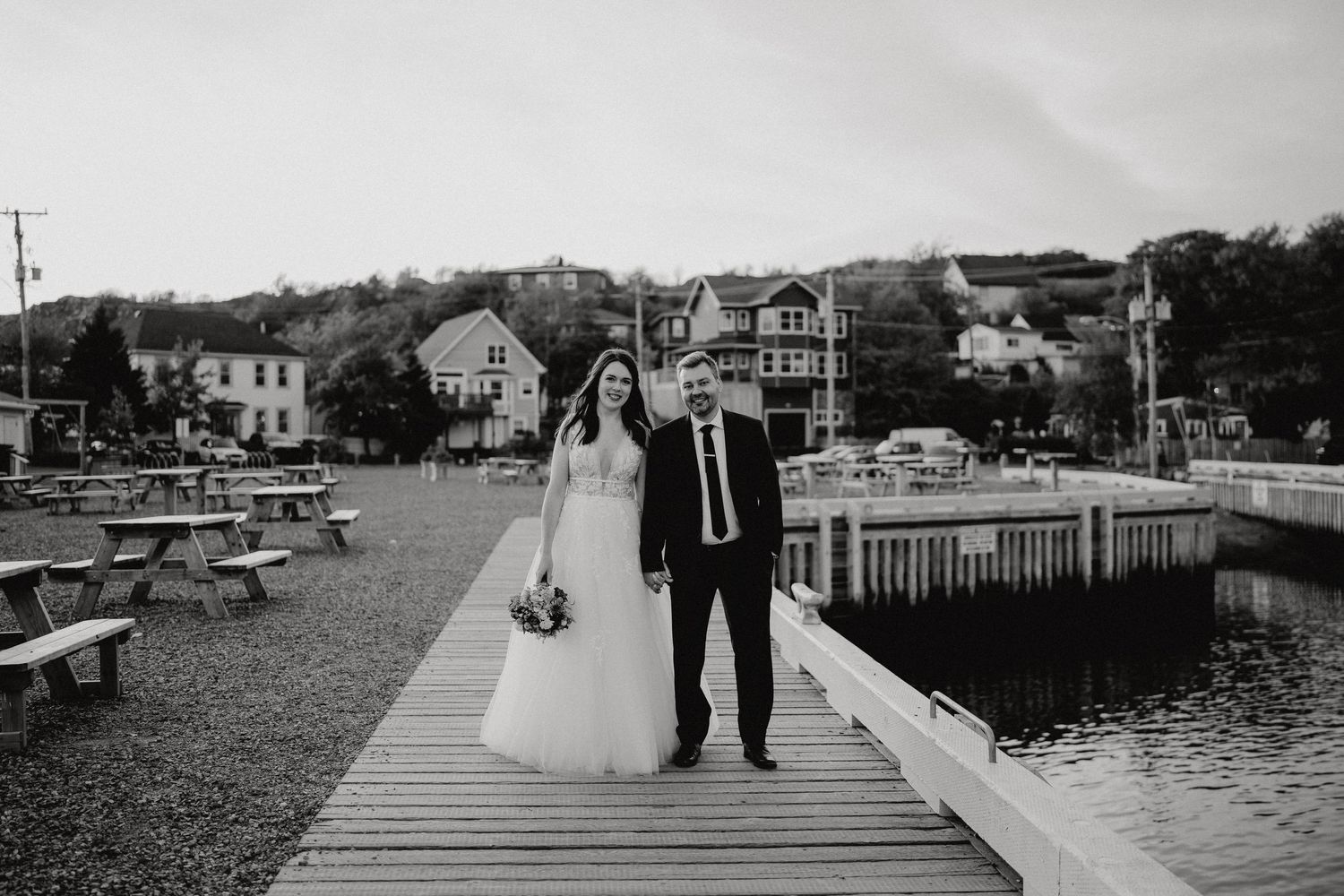 A couple walking along a wooden pier with waterfront homes in the background.