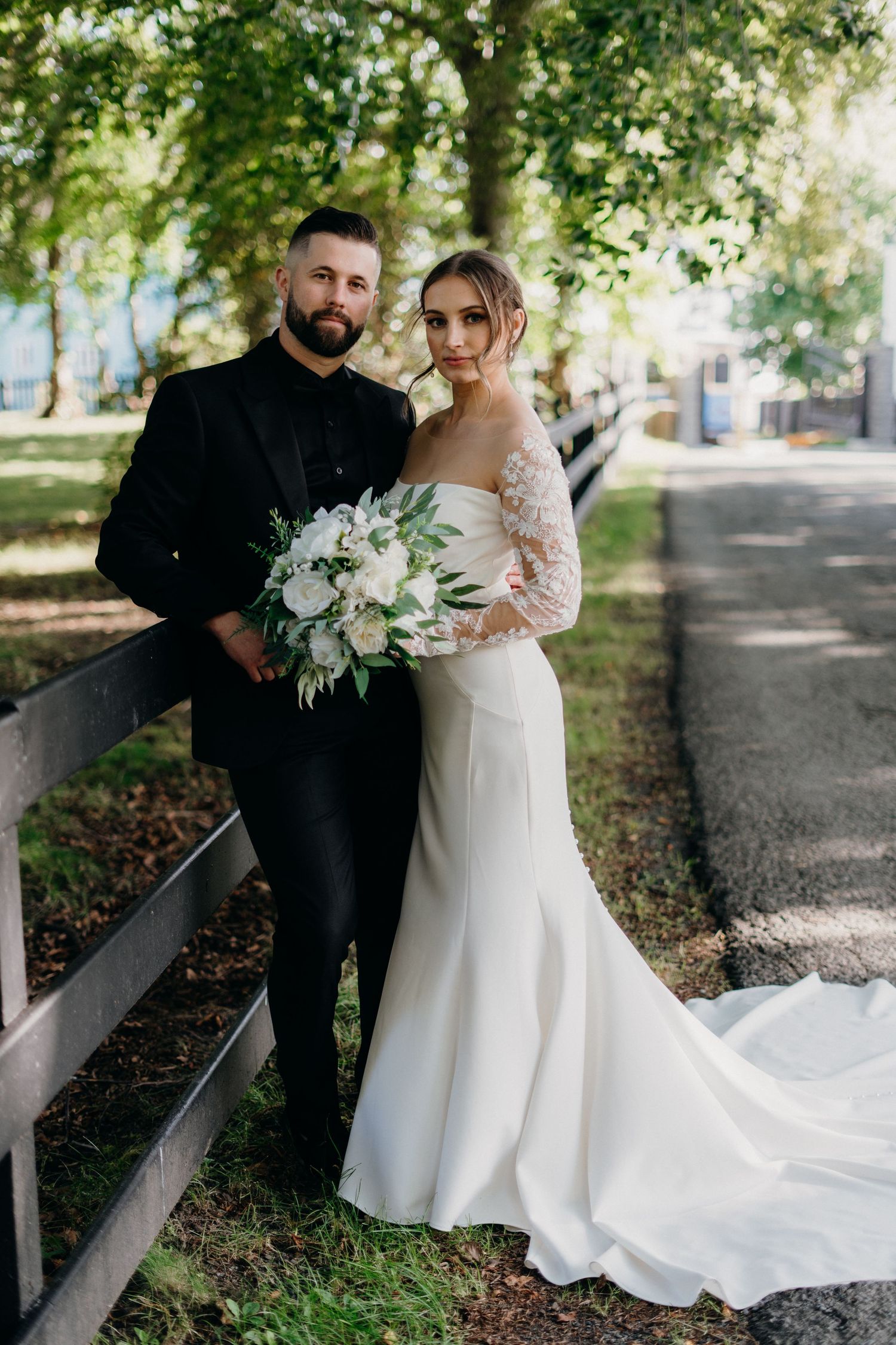 Romantic wedding photos of couple in white dress and black formalwear under tree-lined path with white bouquet.