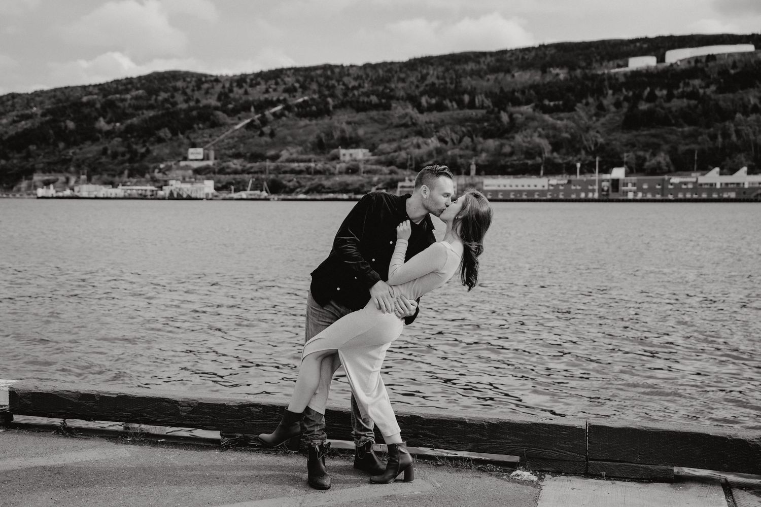 A black and white waterfront engagement photo with mountains and a bridge in the distance.
