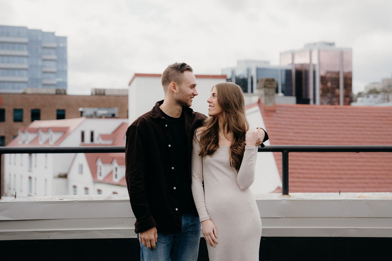 Couple embracing on city rooftop with modern buildings and overcast sky in background.