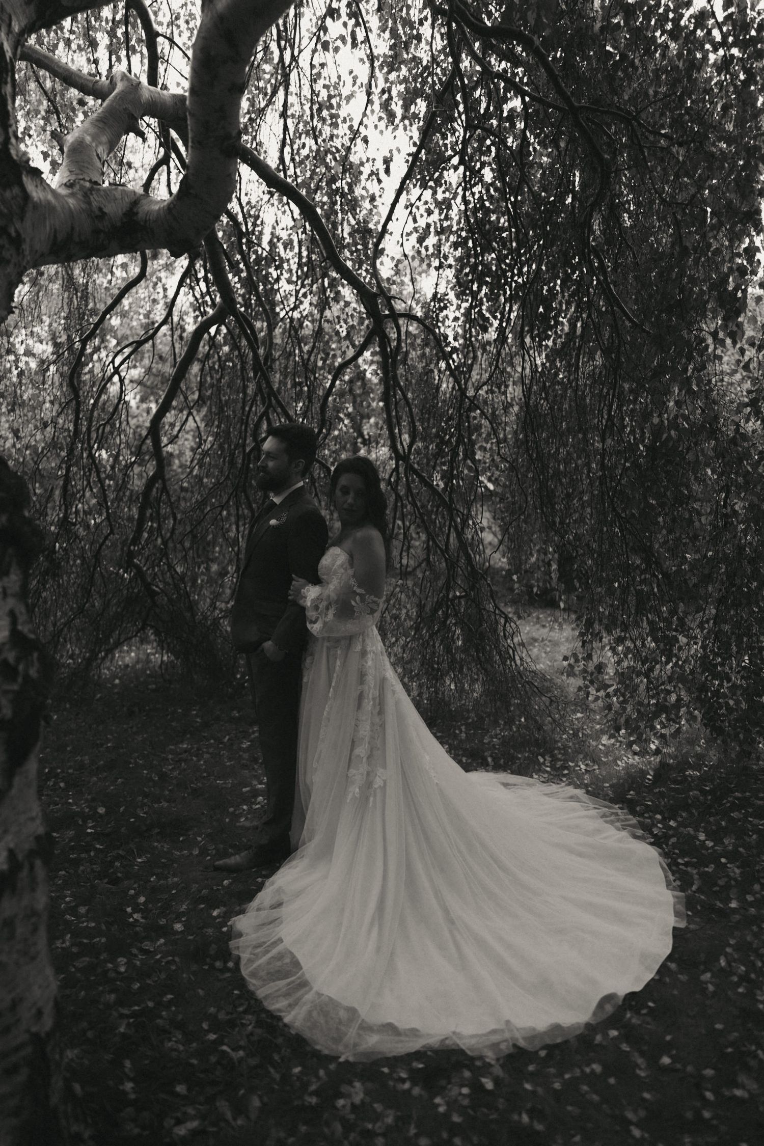 A flowing white wedding gown creates a dramatic silhouette against a moody forest backdrop in black and white photography.