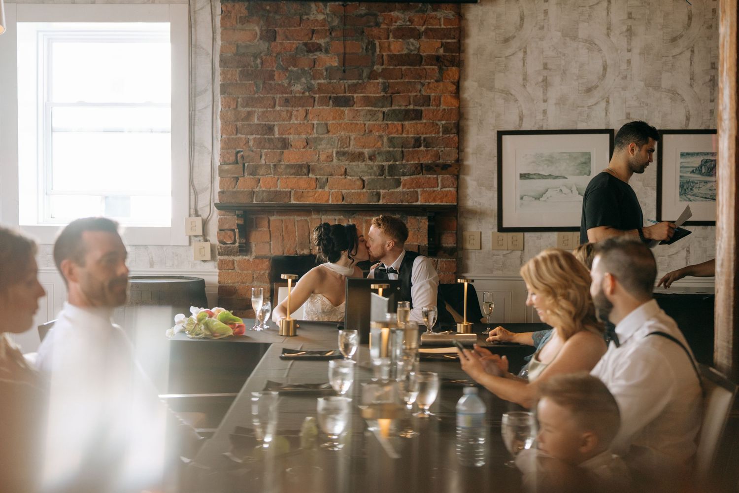 Couple sharing a kiss at a long dining table during a wedding reception in a rustic brick-walled restaurant.