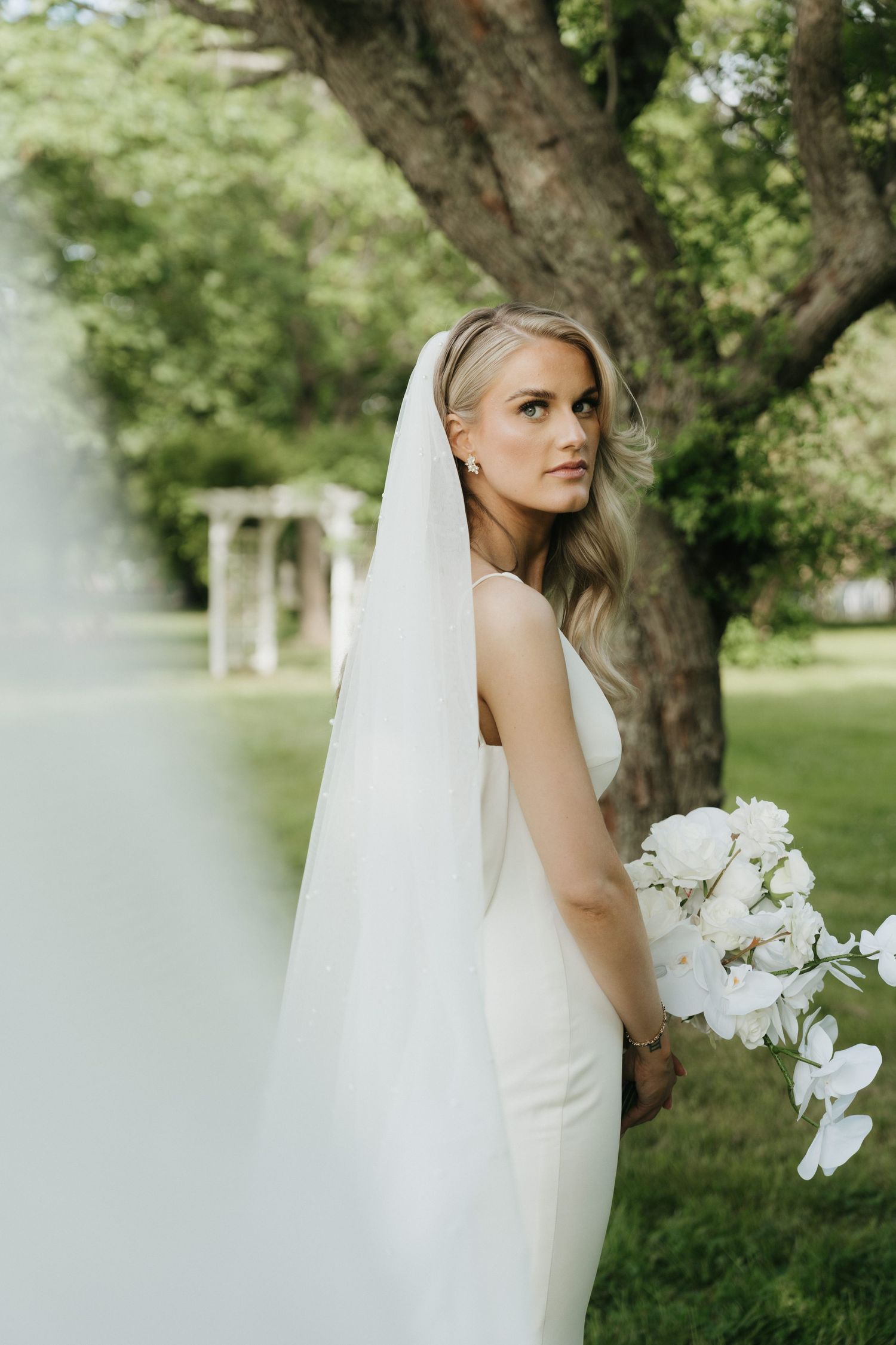 Bride holding white orchids poses in lush garden setting with veil flowing.