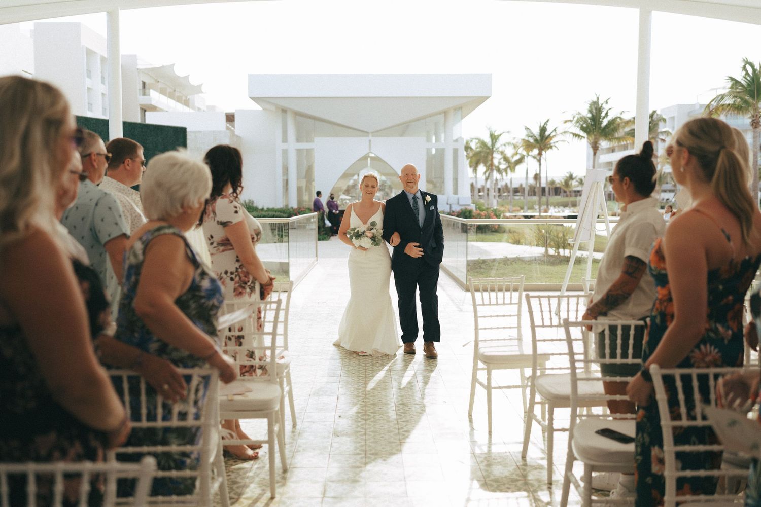 White wedding ceremony space with guests seated in rows facing a beachfront view.