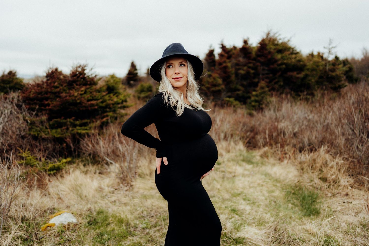 Maternity photo of woman in black dress and hat posing on a coastal trail.