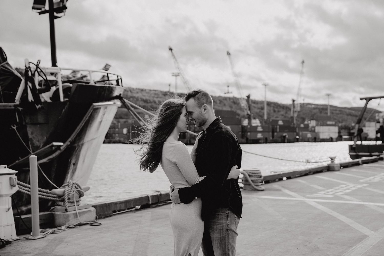 A couple shares a romantic moment on a dock at a harbor under cloudy skies.