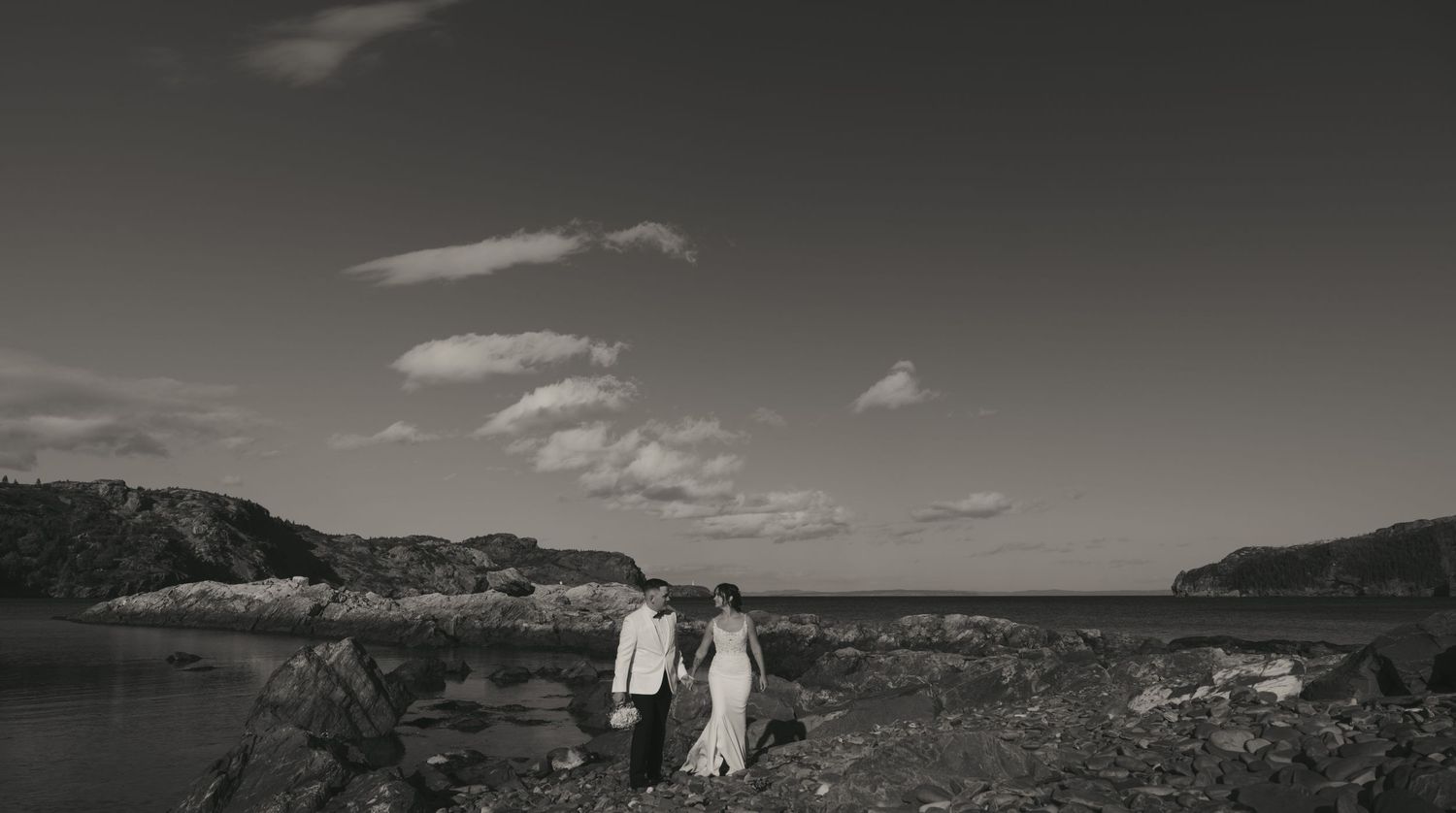 A couple stands together on a rocky coastal landscape under dramatic clouds in black and white.