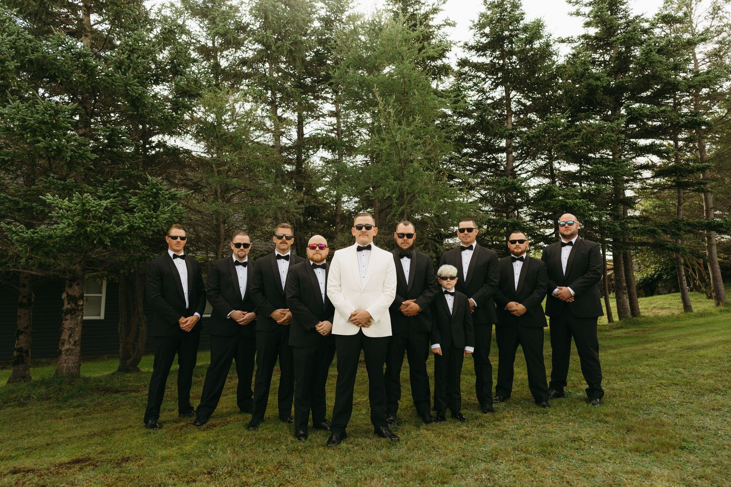 Wedding party in black suits poses together on a green lawn with pine trees in the background.