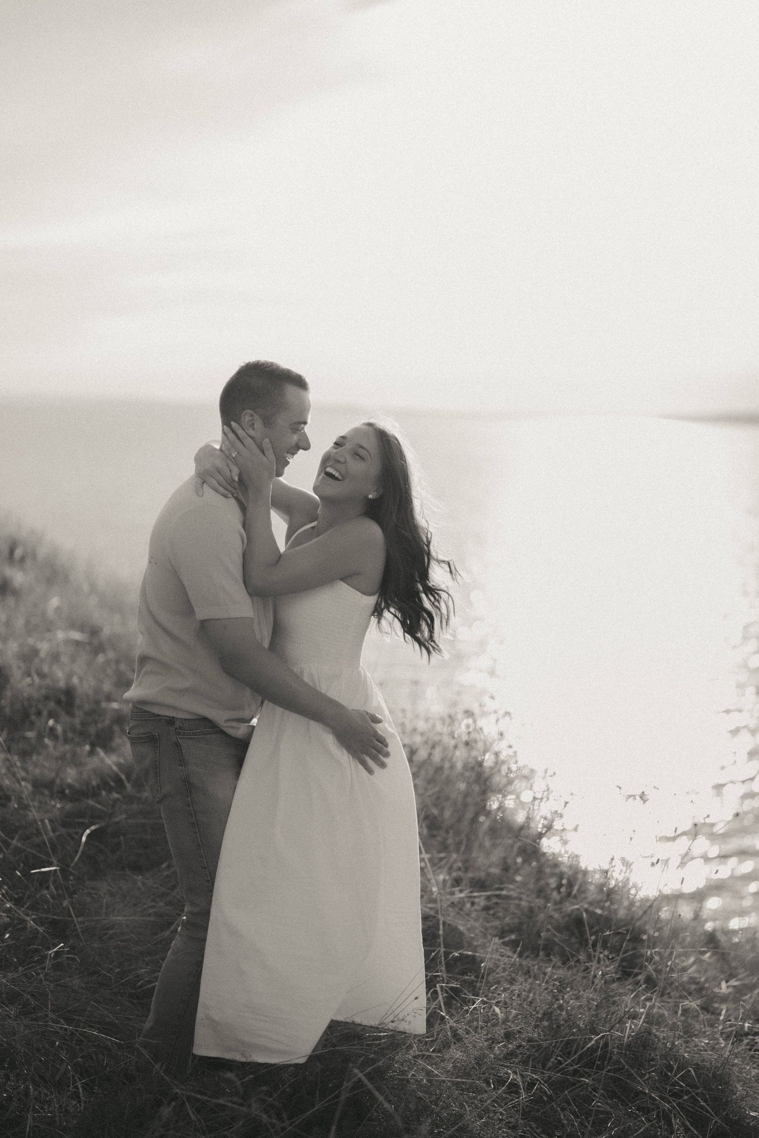 A romantic couple embraces on a hilltop silhouetted against a bright sky in black and white photography.