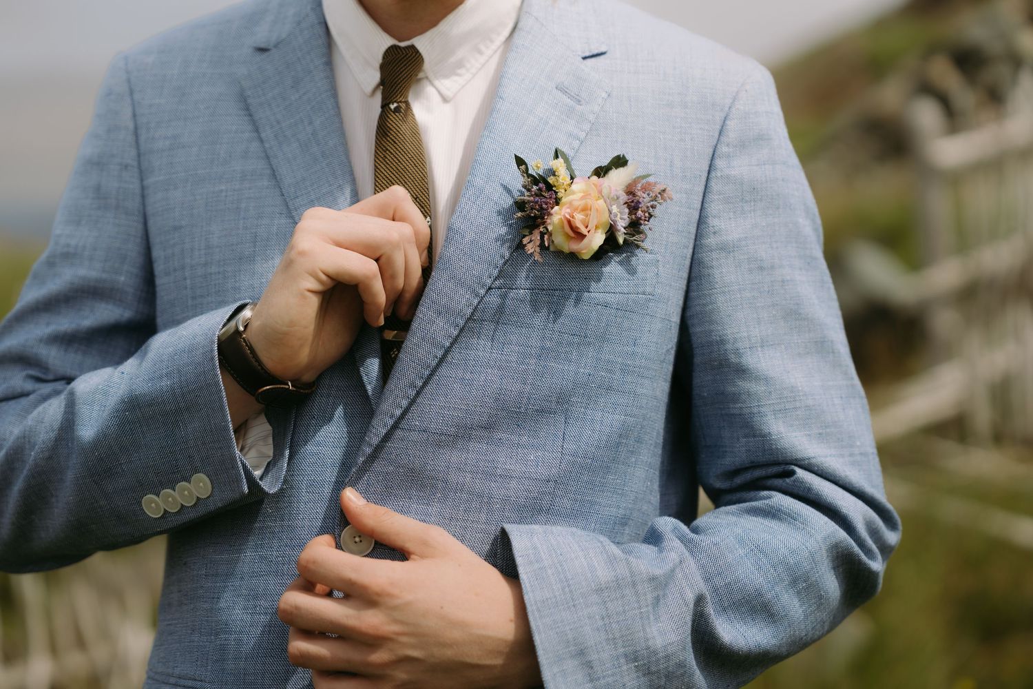 A groom in a light blue suit adjusts his tie while wearing a peach boutonniere at an outdoor wedding.