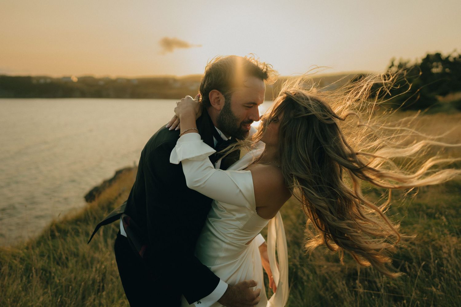 Romantic couple embraces at sunset on a grassy hillside as wind blows through long hair.