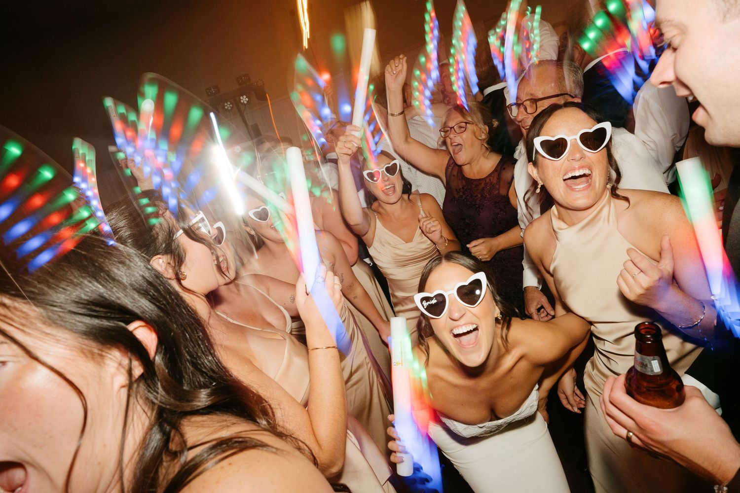 Excited wedding guests dancing with colorful LED glow sticks, wearing heart-shaped sunglasses at a lively reception party.