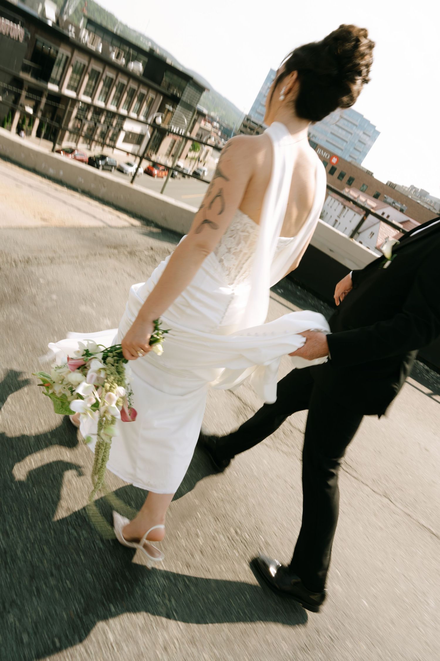 Bride in white halter gown holding bouquet walks with groom in black suit on rooftop after wedding ceremony.