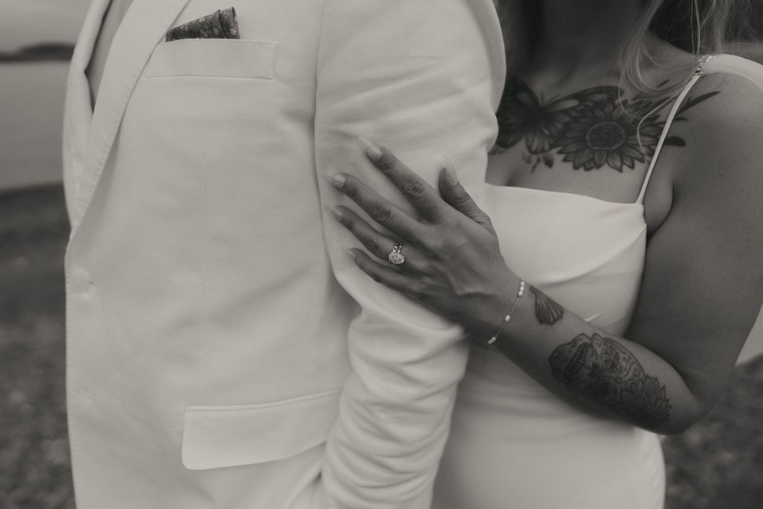 Black and white photo of a tattooed bride embracing a groom in a white suit, showcasing her engagement ring.