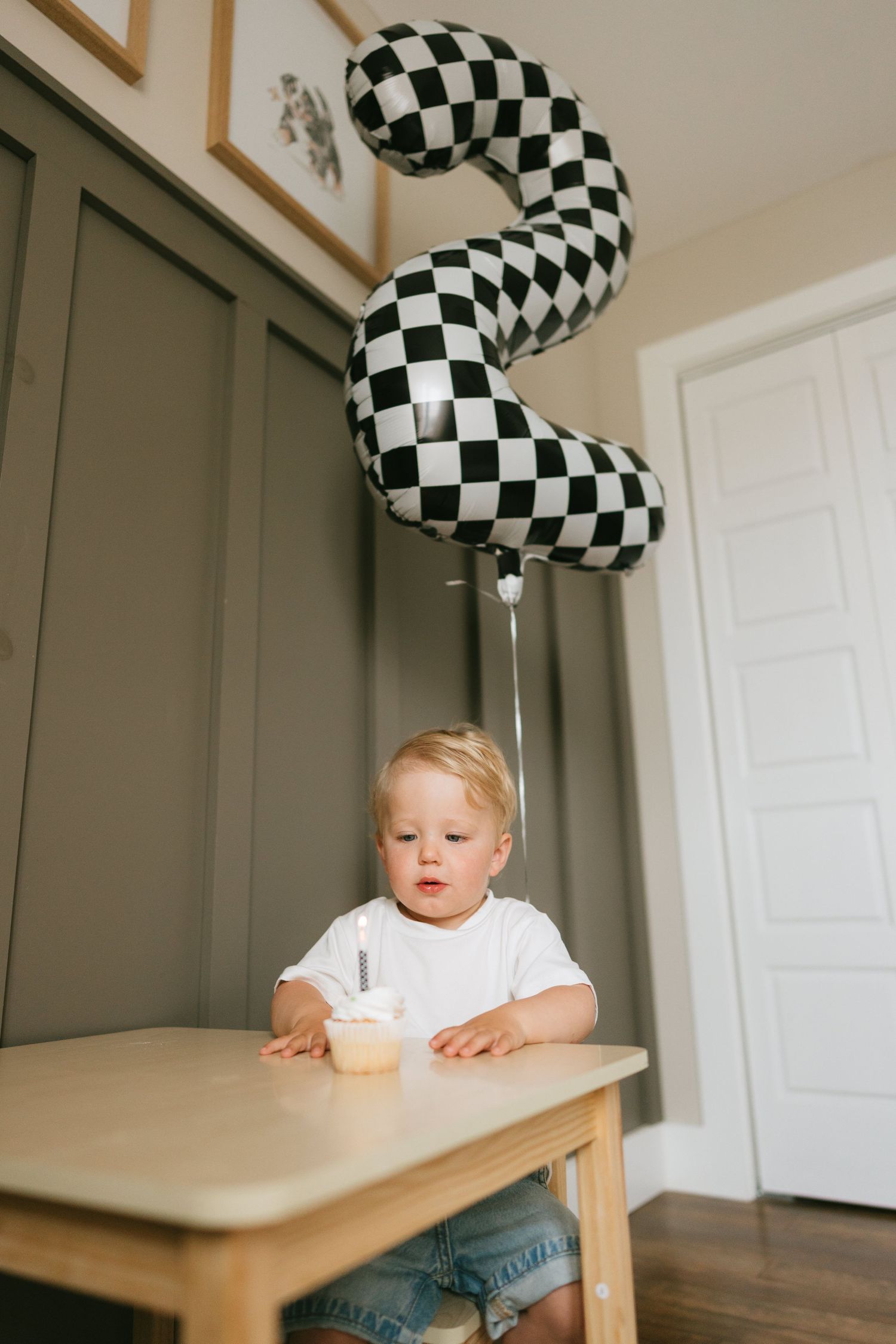 Toddler at wooden table with checkered number 2 balloon celebrating 2nd birthday with cupcake and candle.