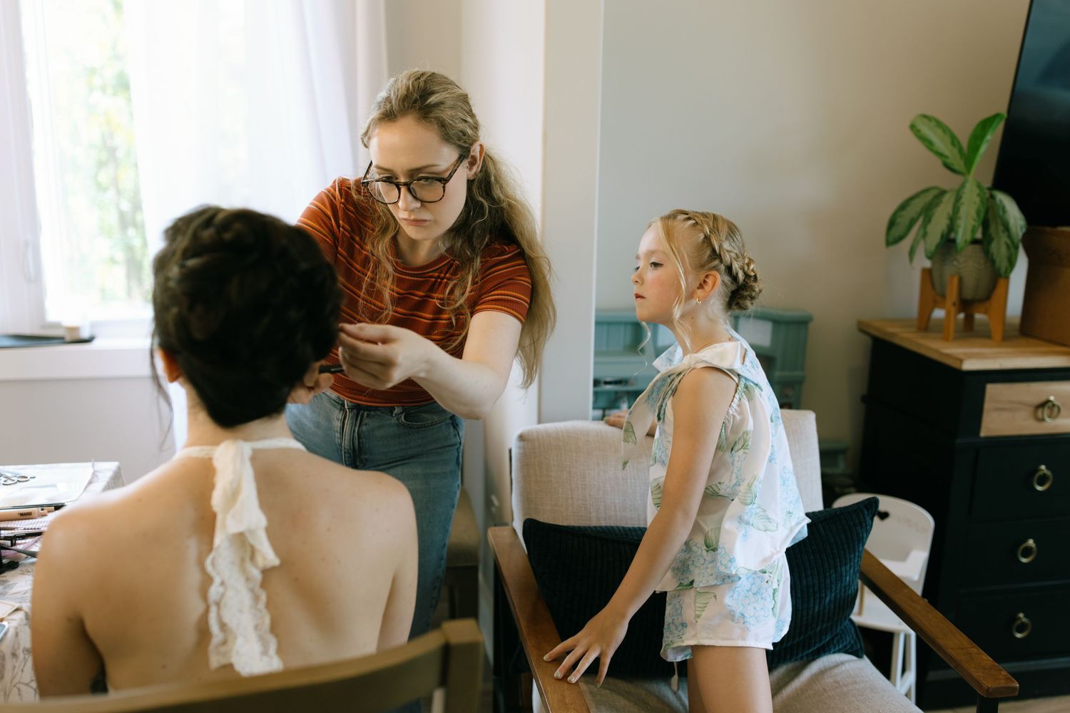 Woman with glasses styles hair of seated person while young girl watches in a bright home interior.