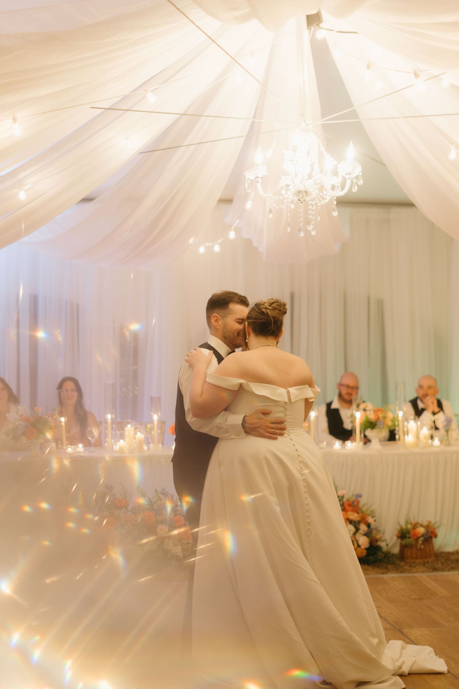 A couple dances beneath elegant white draping and chandeliers in a beautifully lit reception space.
