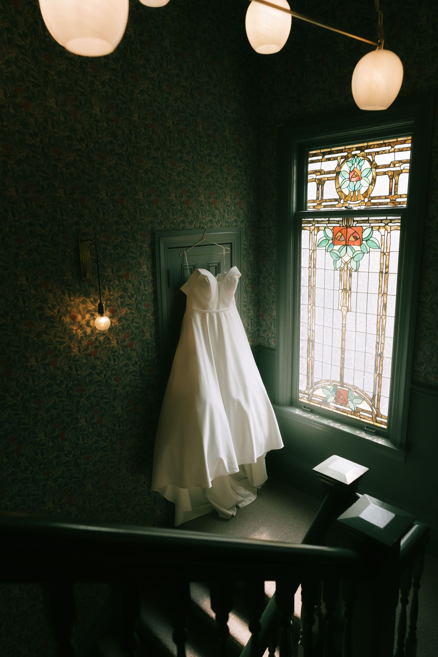 White wedding dress hanging by stained glass window in dark moody room.