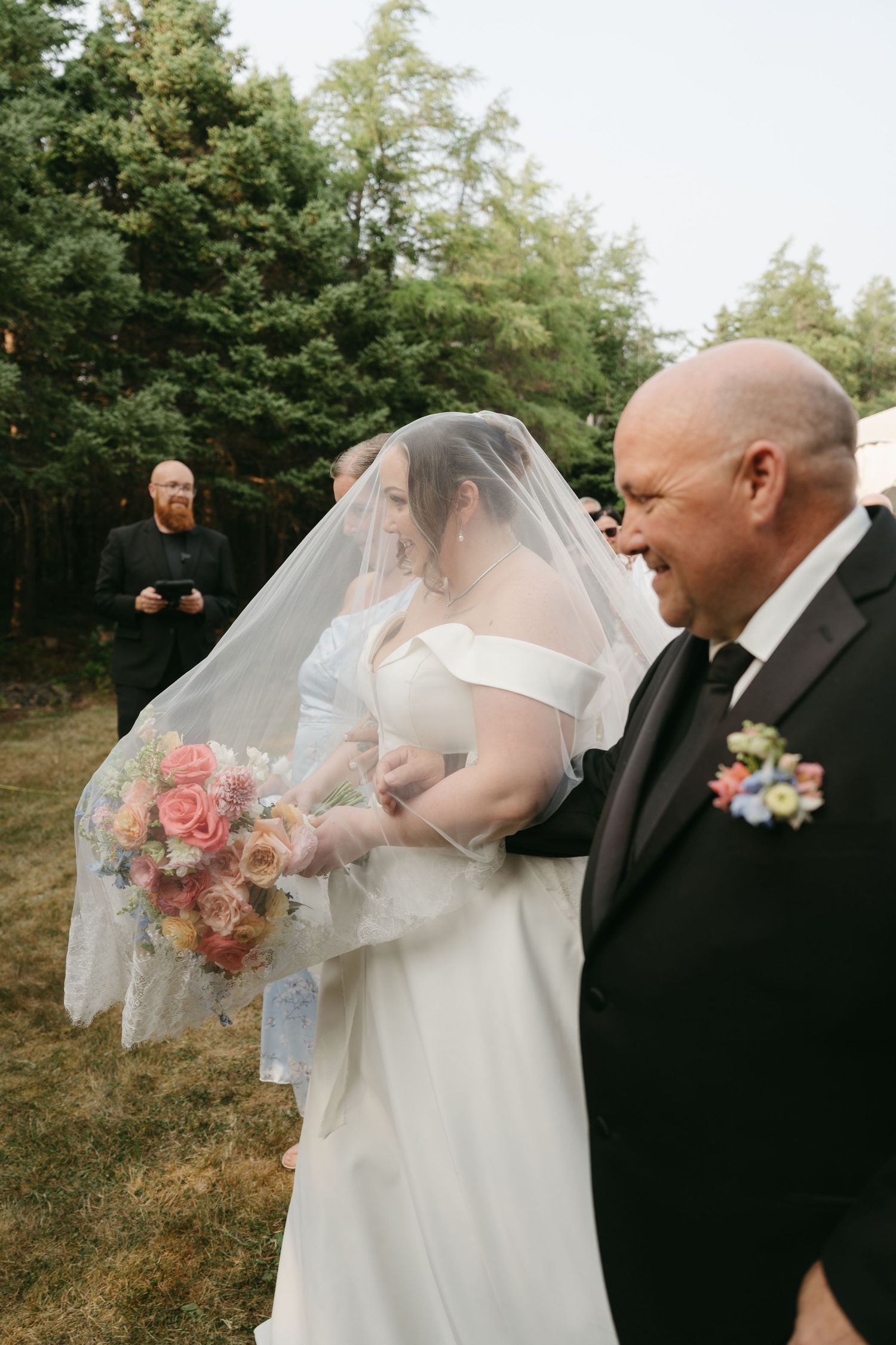 Intimate outdoor wedding moment showing a tender interaction through a bridal veil in front of evergreen trees.