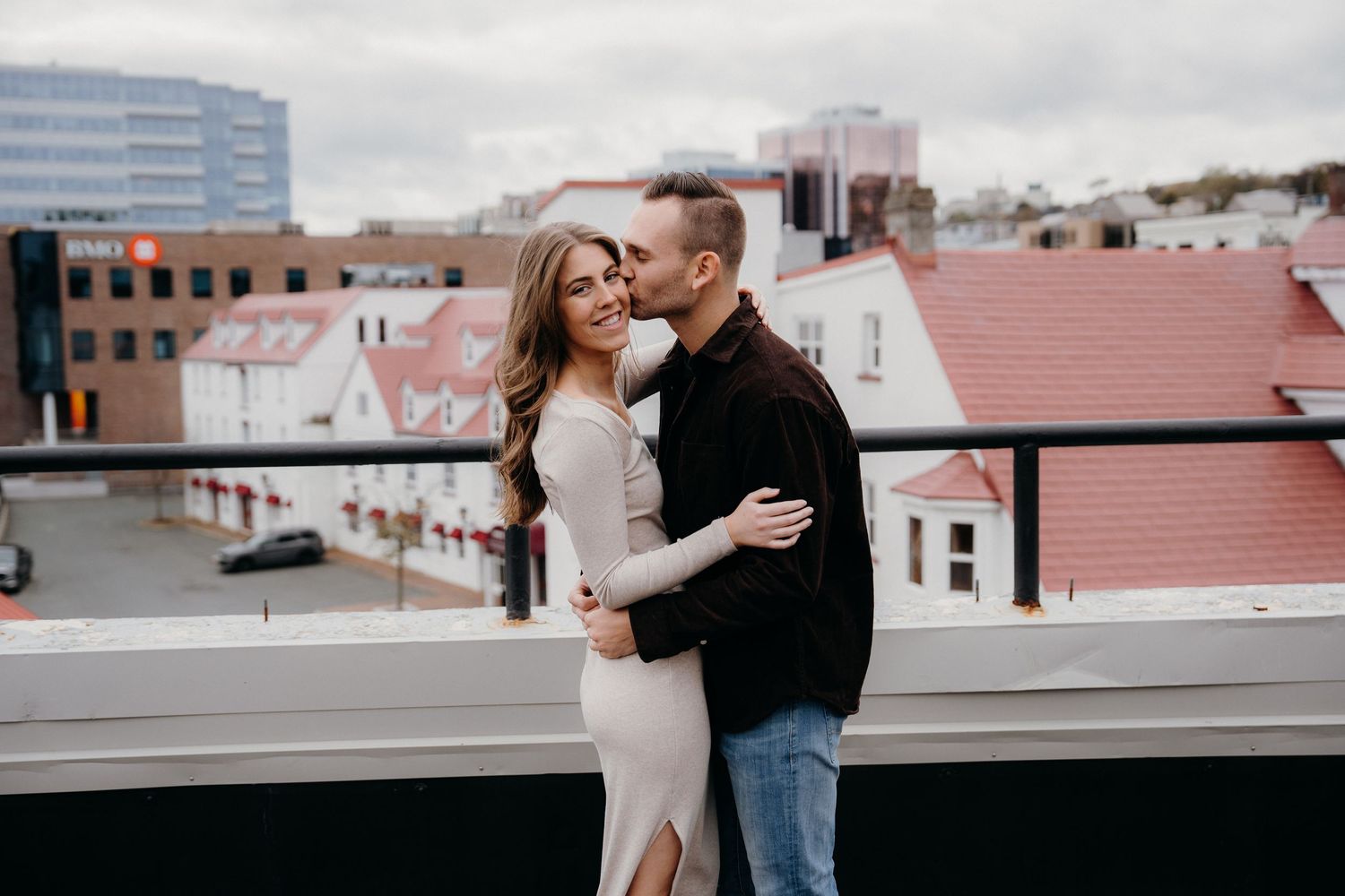 A romantic couple shares a kiss on an urban rooftop with city buildings behind them.