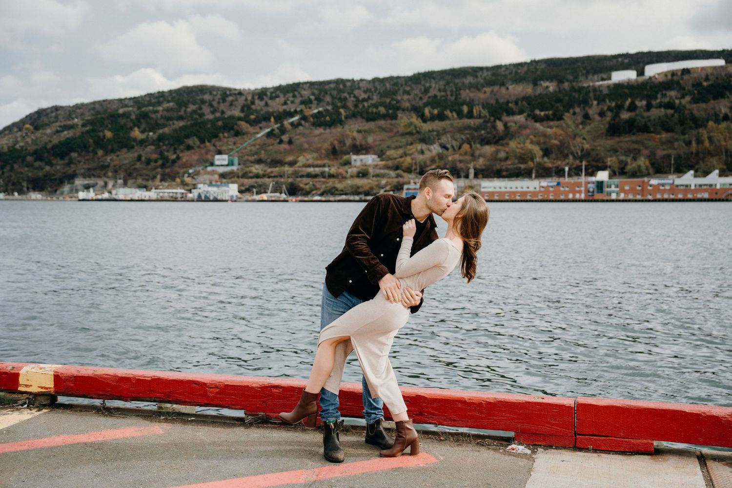 A couple shares an intimate moment along a red-painted waterfront wall with water and hills in the background.