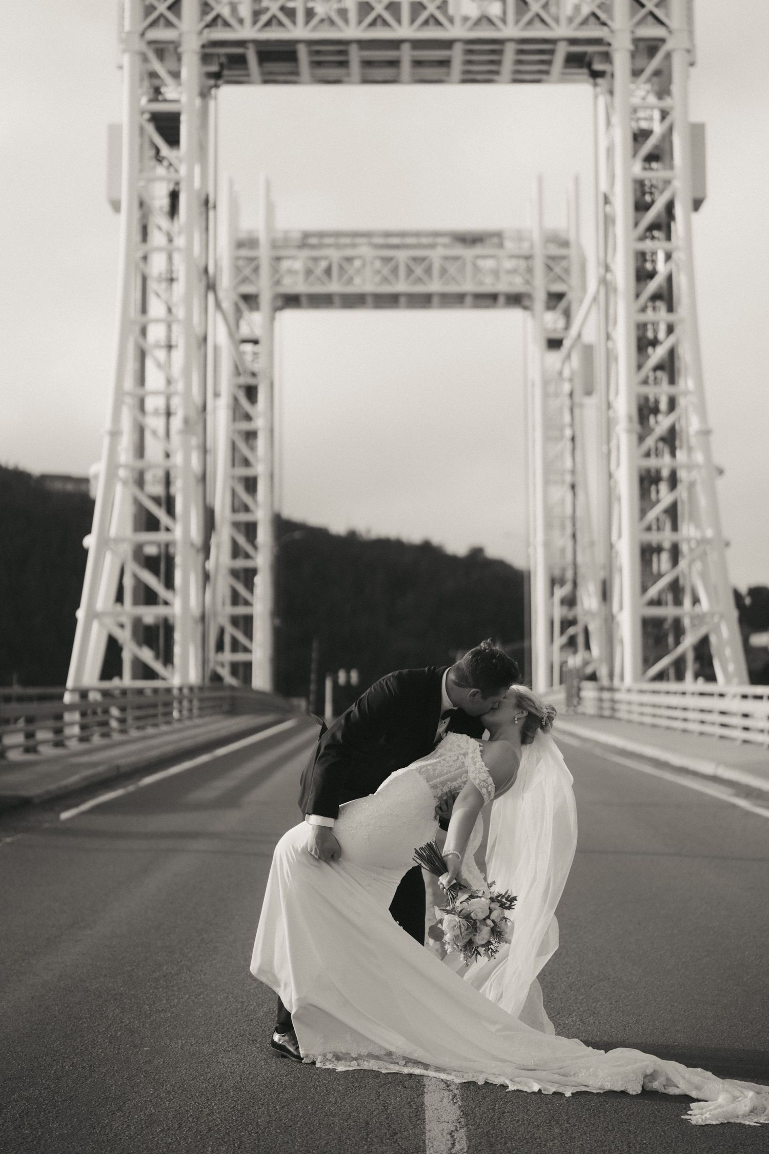 Black and white photo of bride and groom sharing a romantic dip kiss on a bridge road, with towering steel structure behind them.