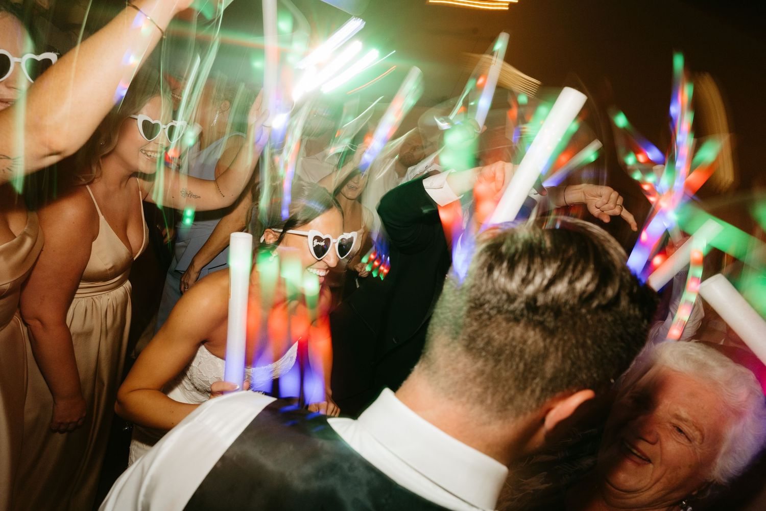 Wedding guests dancing with colorful LED glow sticks at a lively reception party celebration at night.