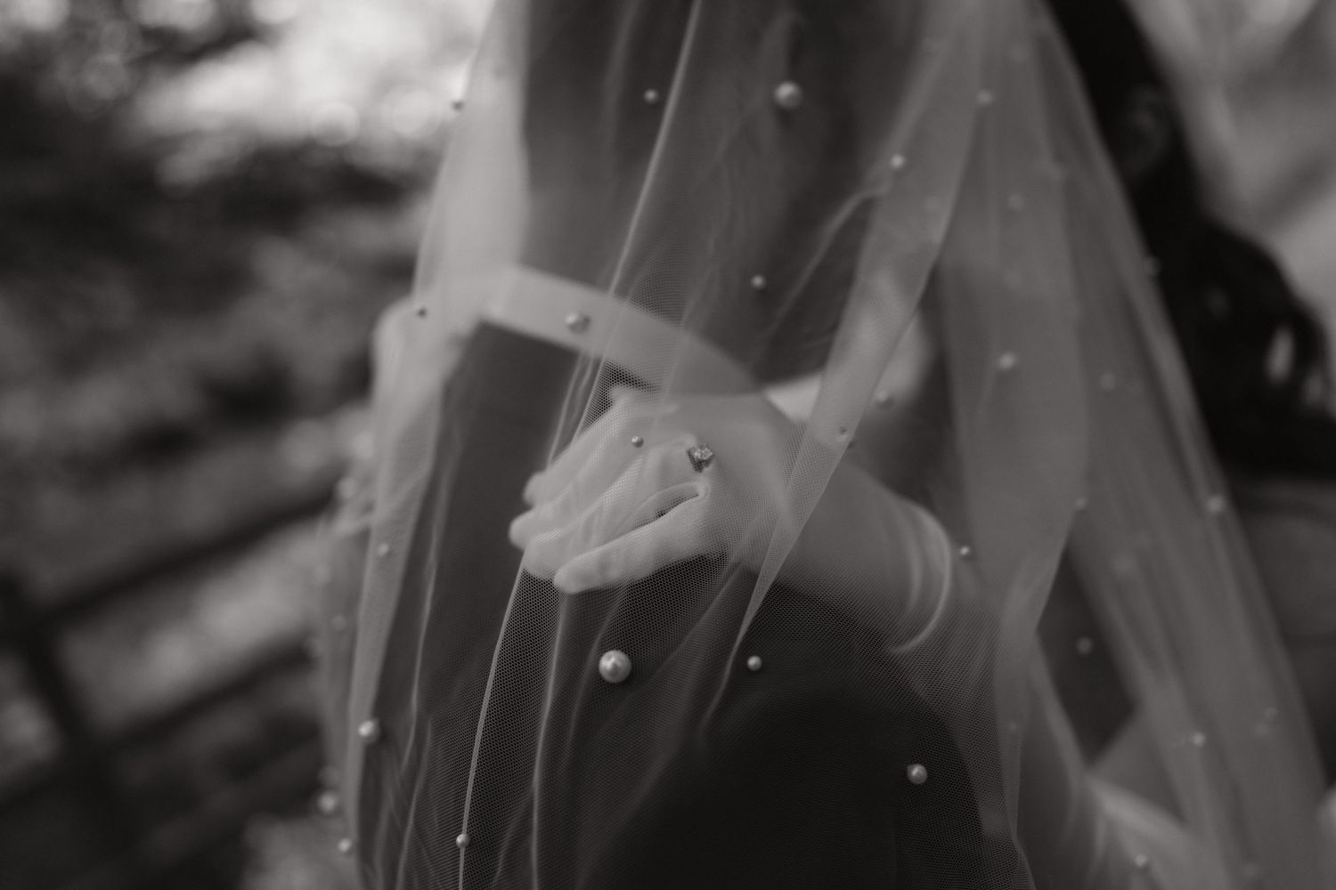 Black and white photo of bride holding wedding veil with pearl details, wearing engagement ring.
