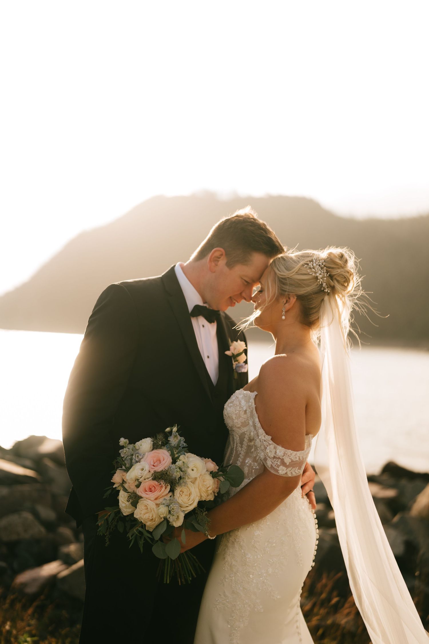 Bride and groom share romantic forehead touch at golden hour by rocky waterfront, bride holding blush bouquet in lace gown.