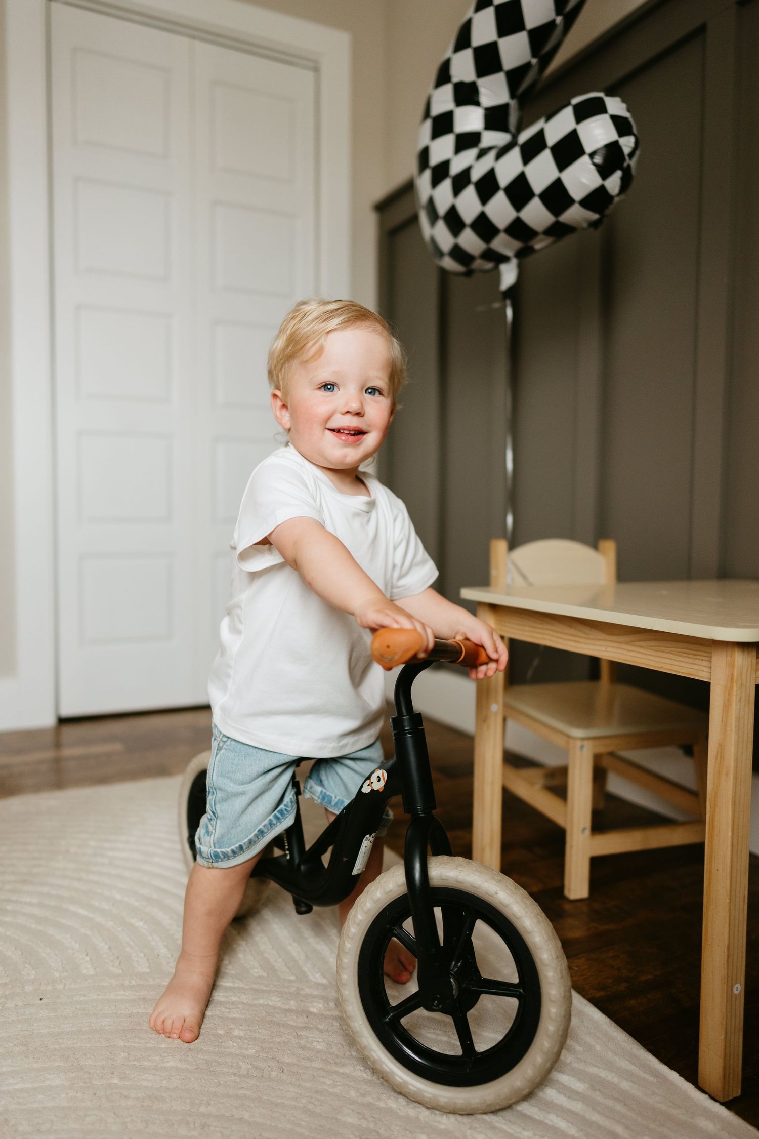 Smiling toddler with blonde hair rides a balance bike indoors near a wooden table with checkered balloons.