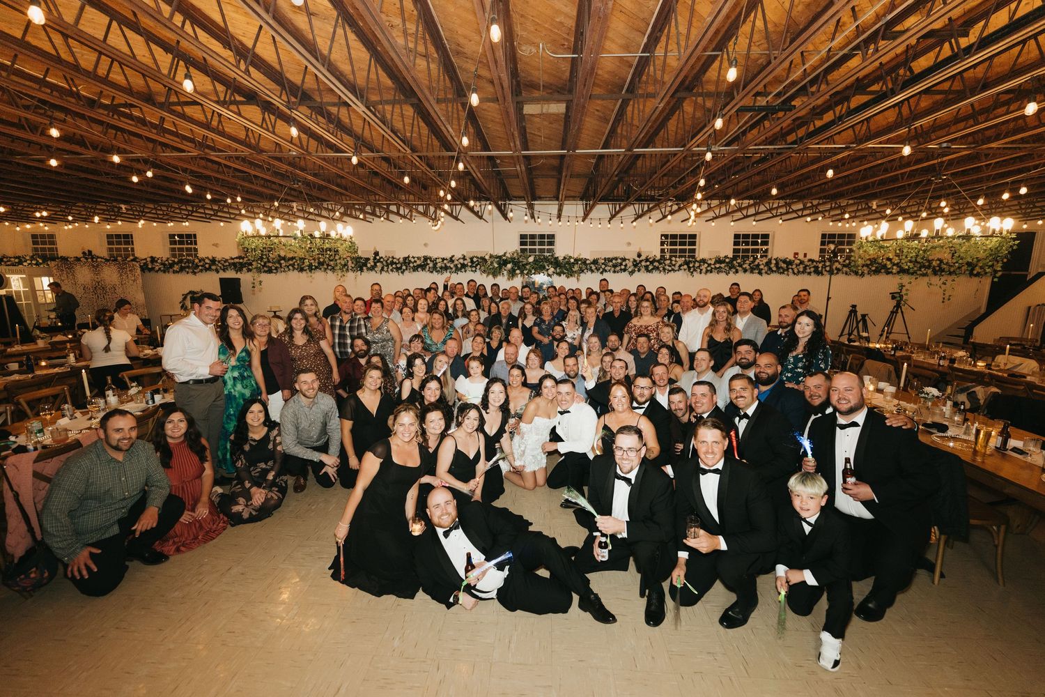 A panoramic view of a large indoor wedding reception venue with wooden beams and string lights overhead.