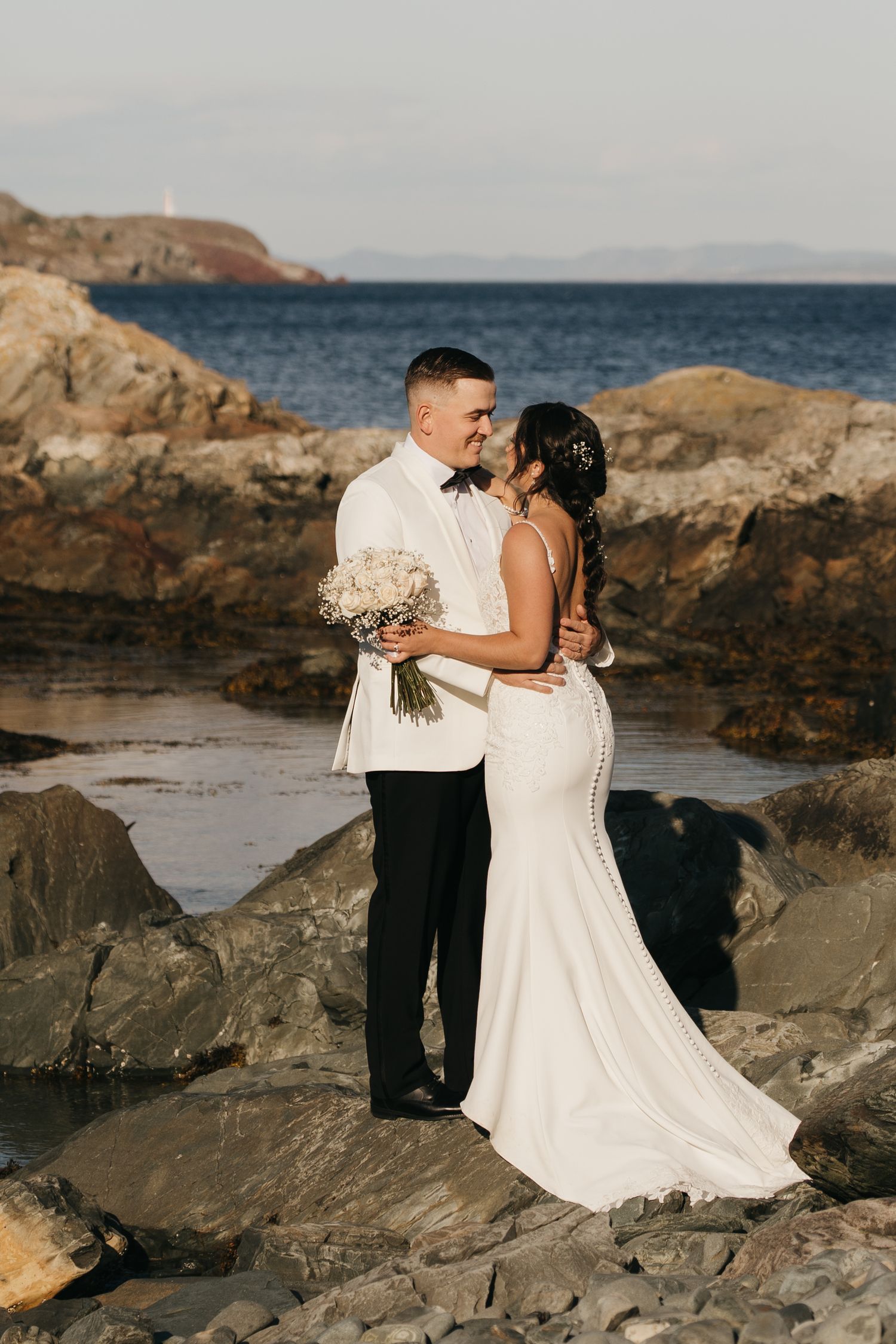 A couple shares an intimate moment on rocky coastal cliffs at sunset, with the bride holding a white floral bouquet.