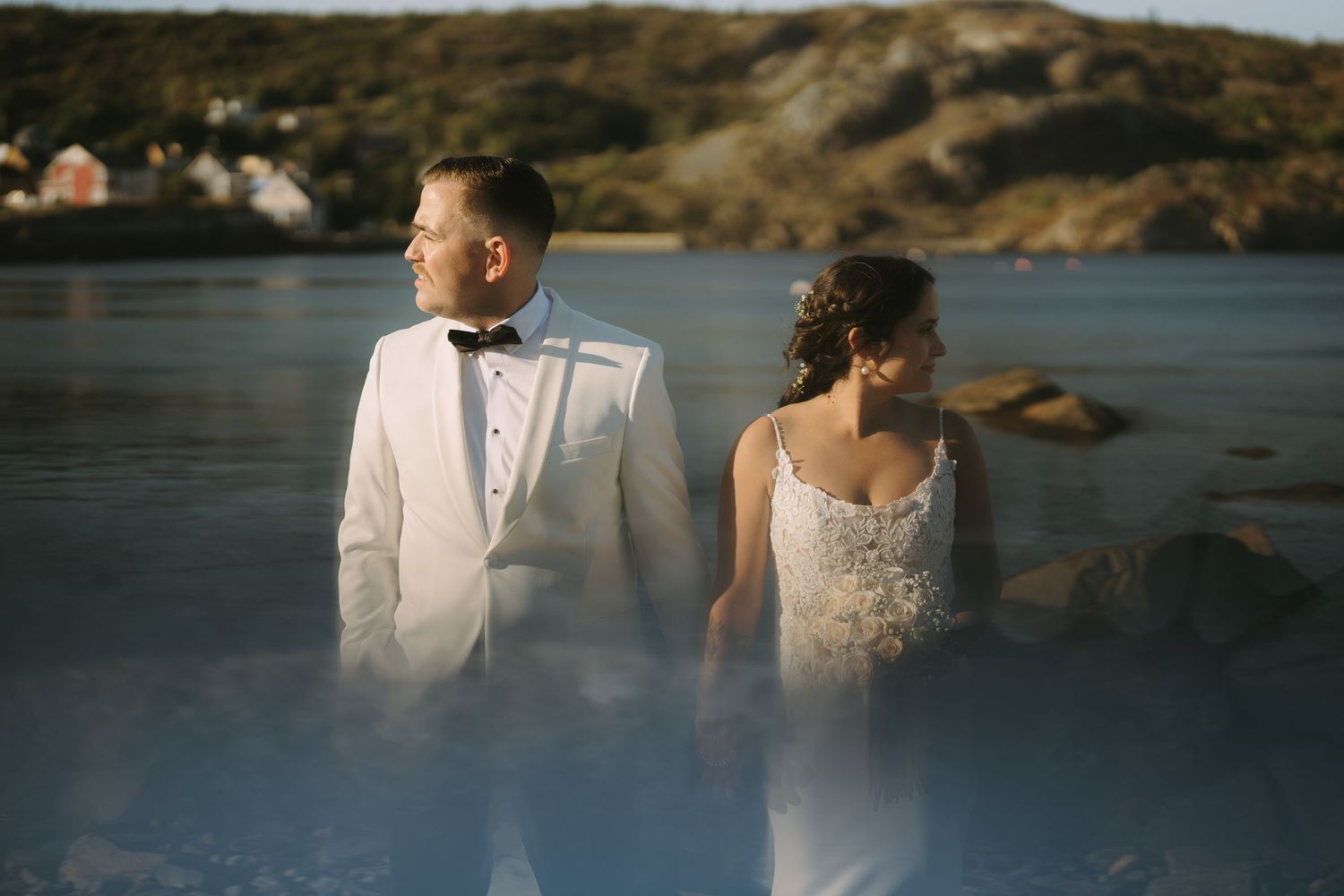 A couple in wedding attire stands together on a rocky coastline at sunset with ocean waves in the background.