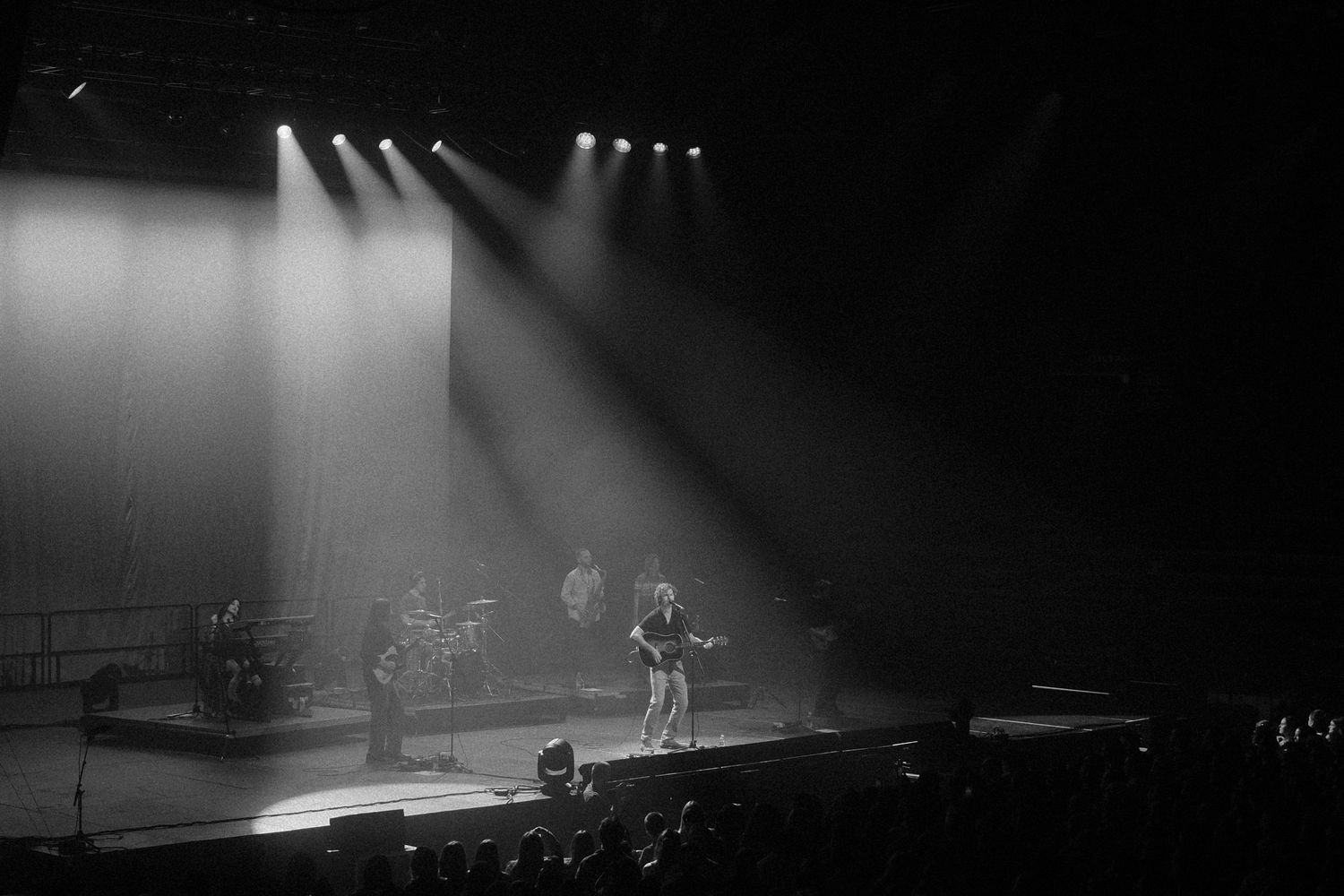Musicians perform on a dimly lit concert stage with dramatic spotlights beaming down through atmospheric haze.