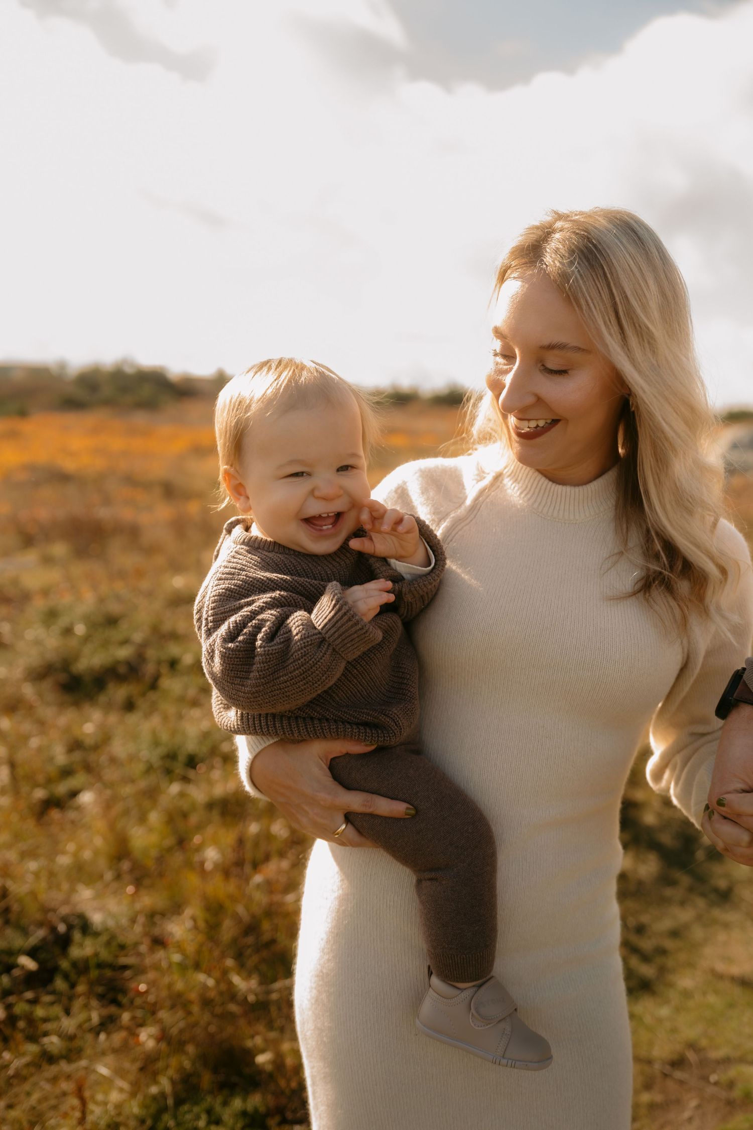A joyful moment captured in golden sunlight as a woman in white holds a baby wearing a brown sweater in a field.