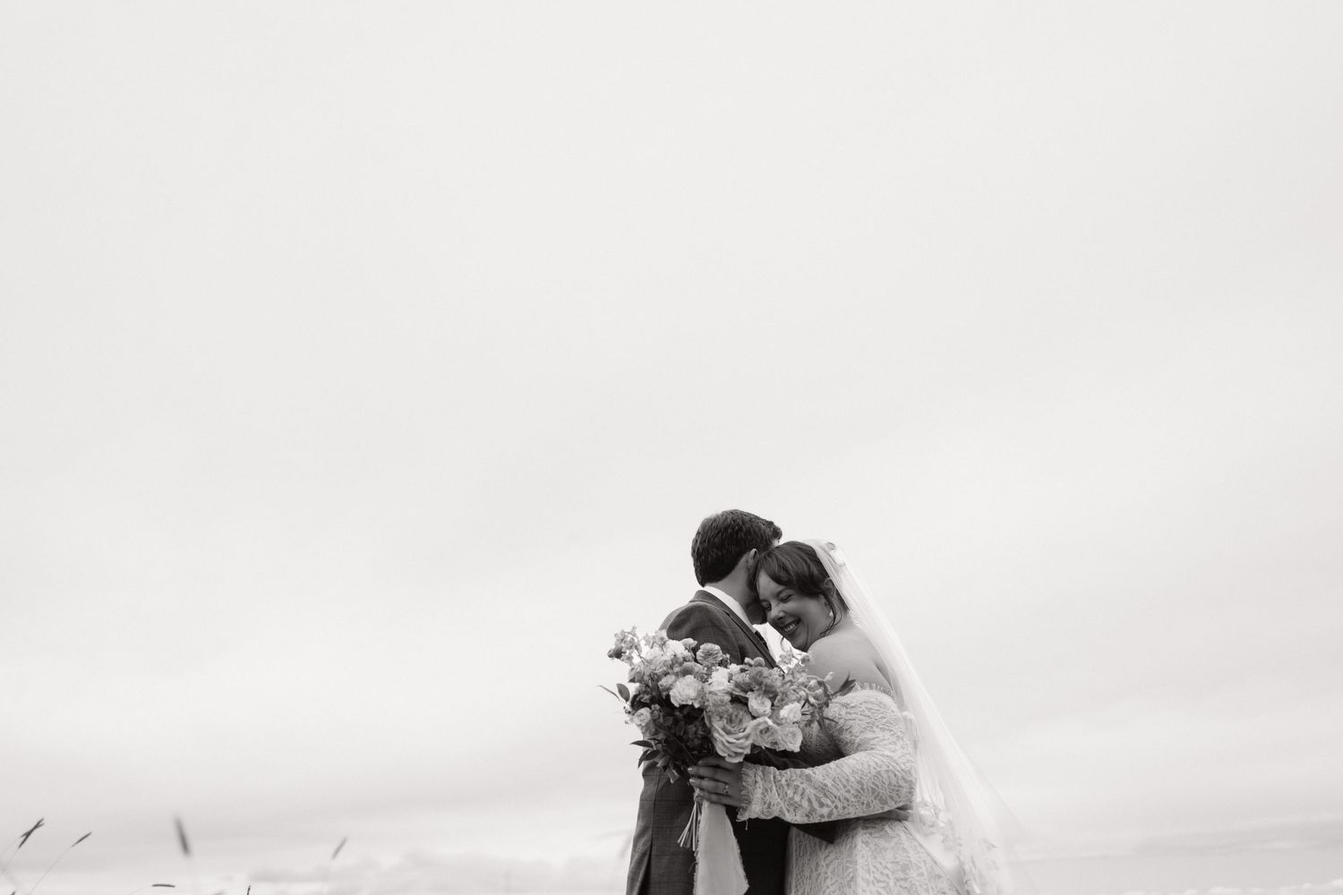 Black and white photo of a bride and groom embracing tenderly outdoors, holding a bouquet on their wedding day.