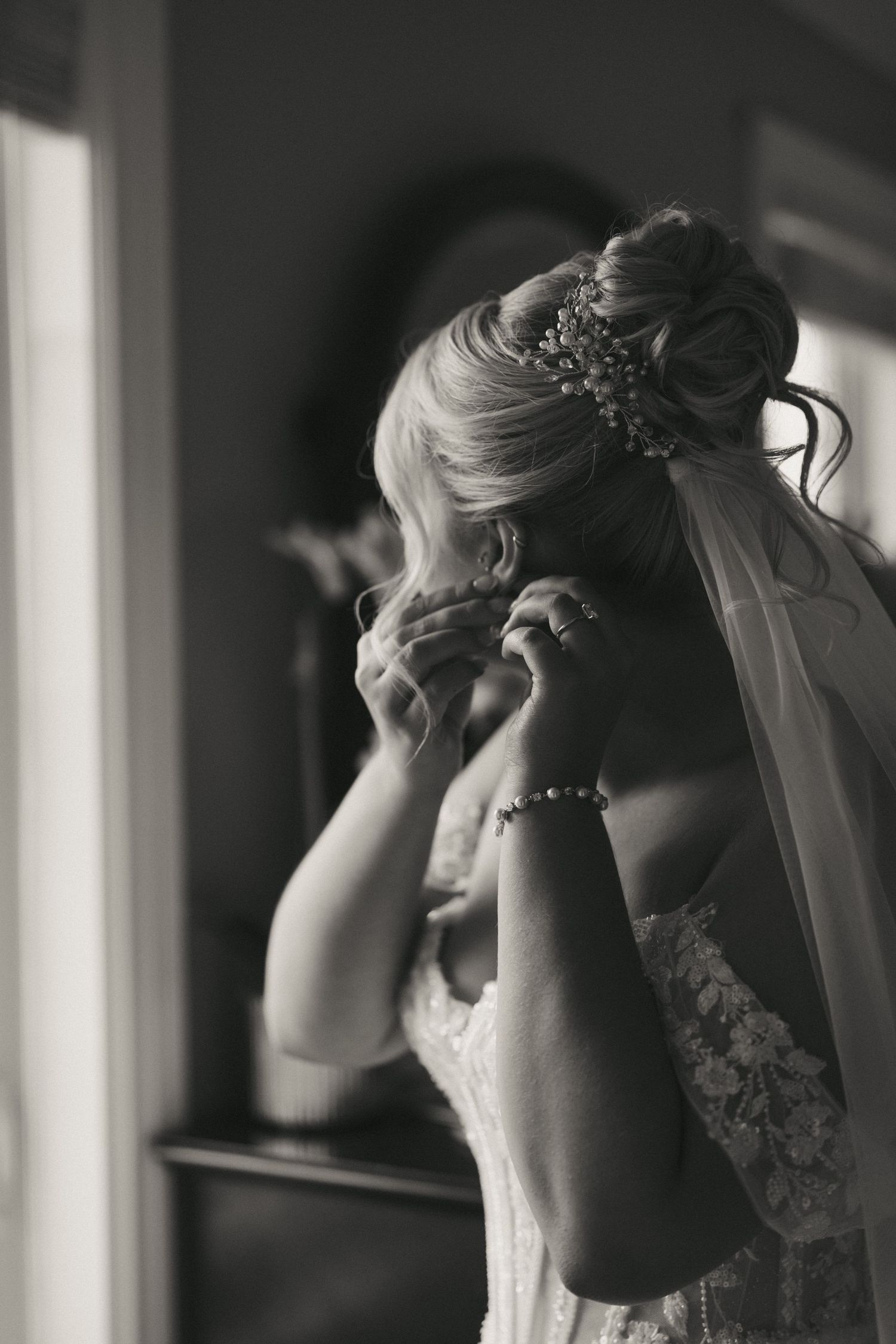 Black and white photo of a bride in lace gown adjusting her earring, wearing a veil and hair comb.