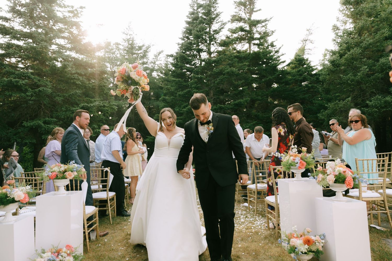 Forest wedding ceremony with guests seated on gold chairs and floral decorations.