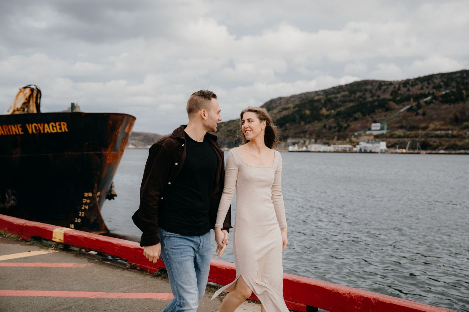 Two people walking along a red-lined dock edge with water and mountains in the background.
