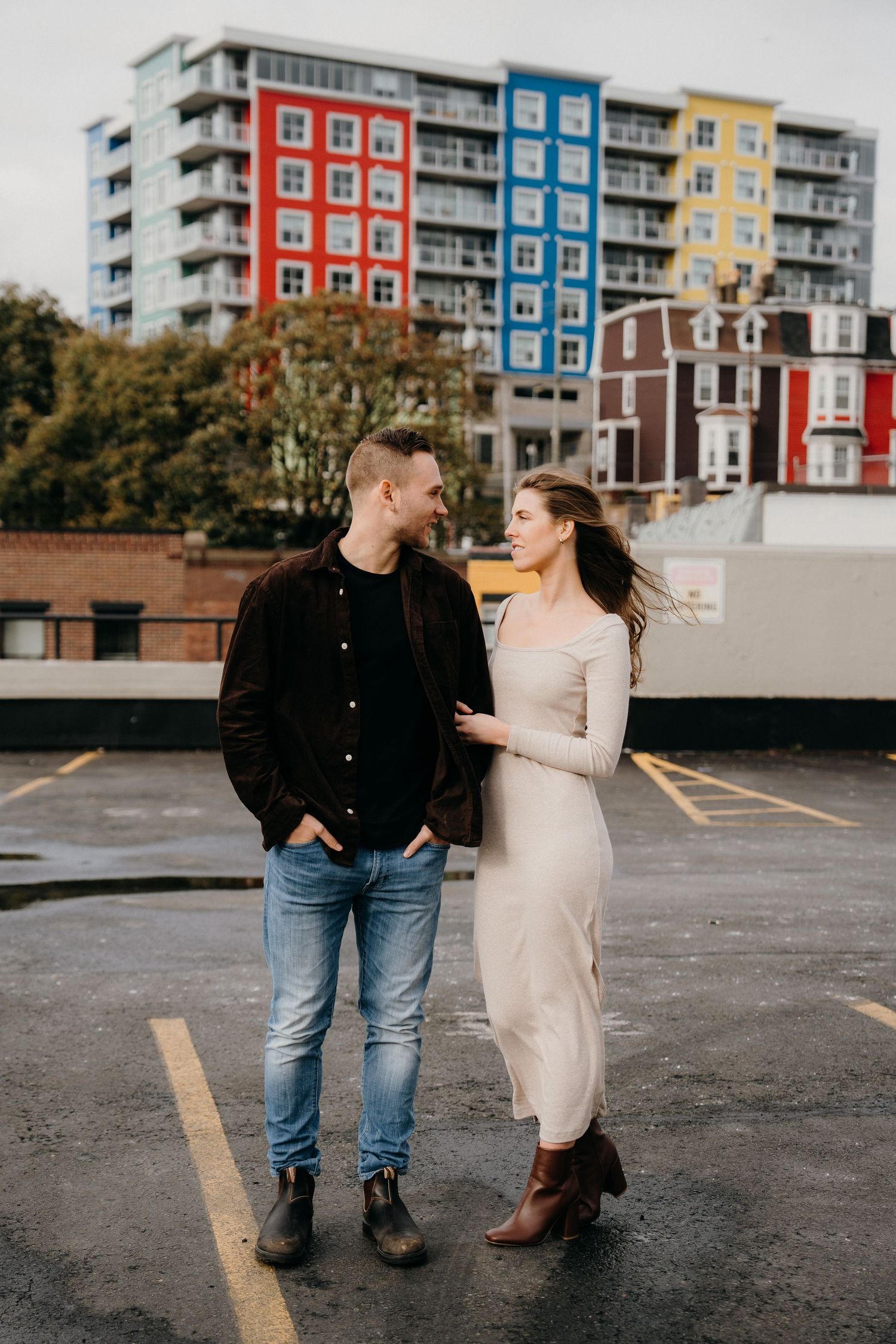 A couple in casual attire walks together on a wet parking lot surface near modern apartments.