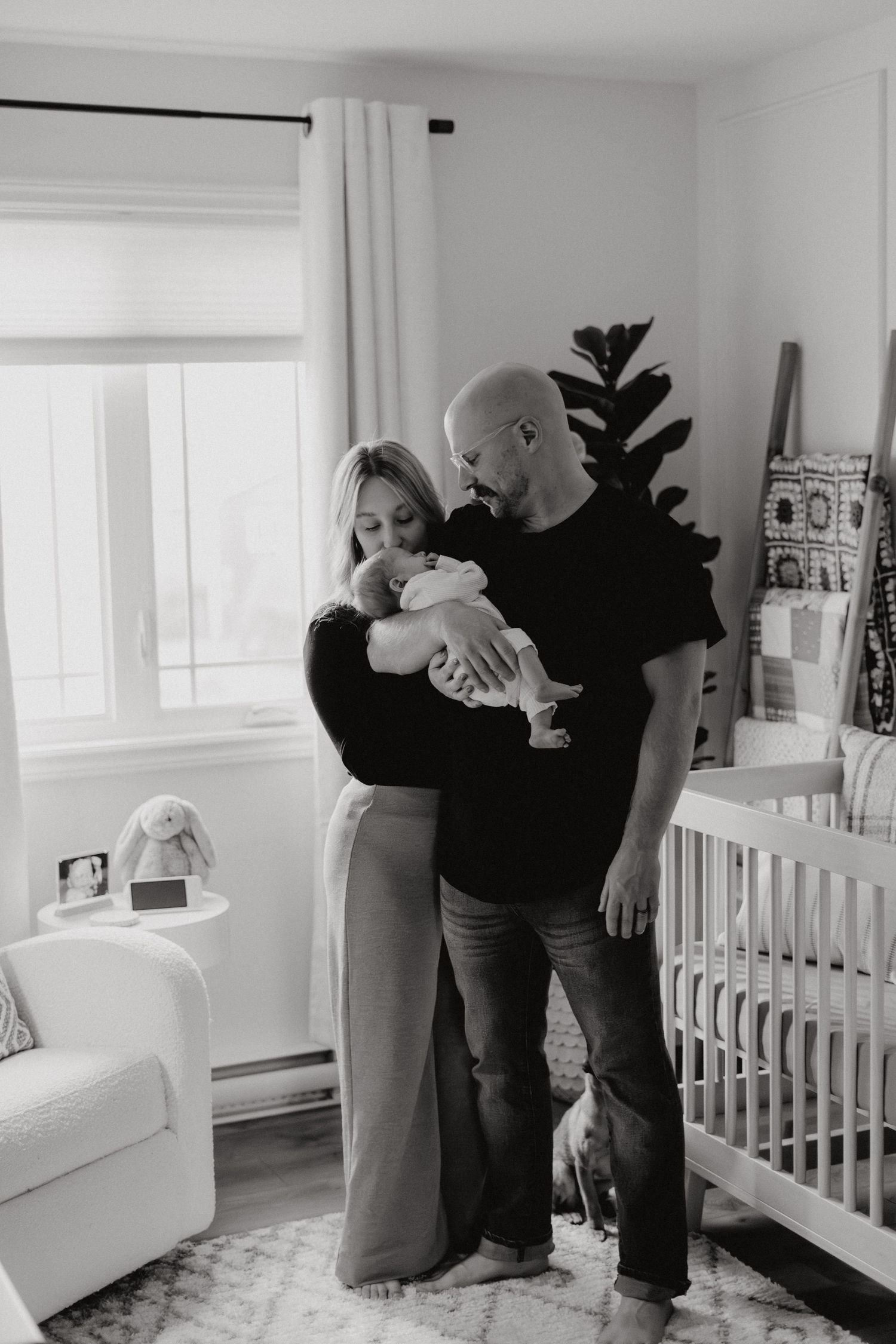 Tender black and white family moment captured in front of a white crib in a nursery.