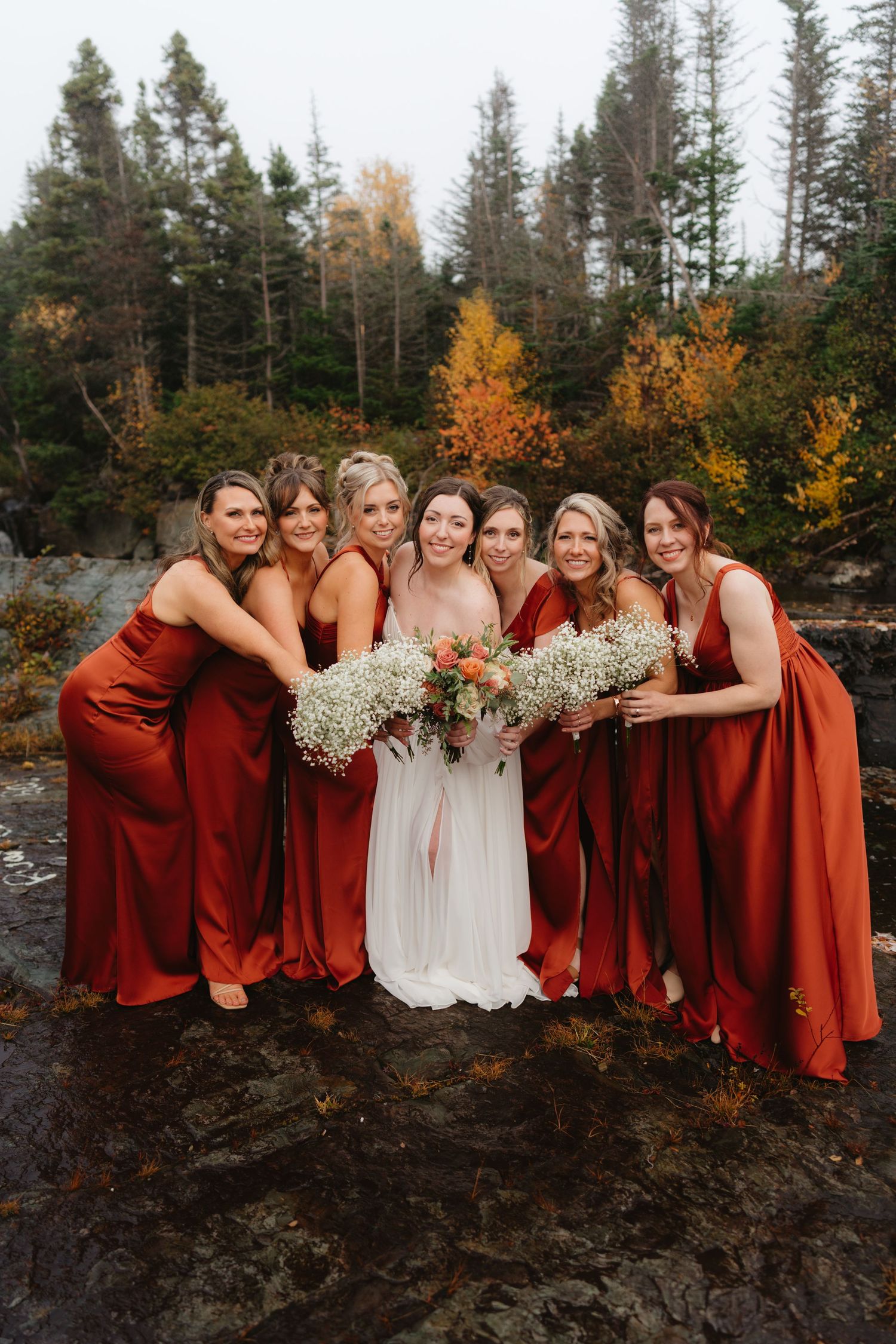 Wedding party in burgundy dresses poses together against autumn foliage and evergreen trees in outdoor setting.